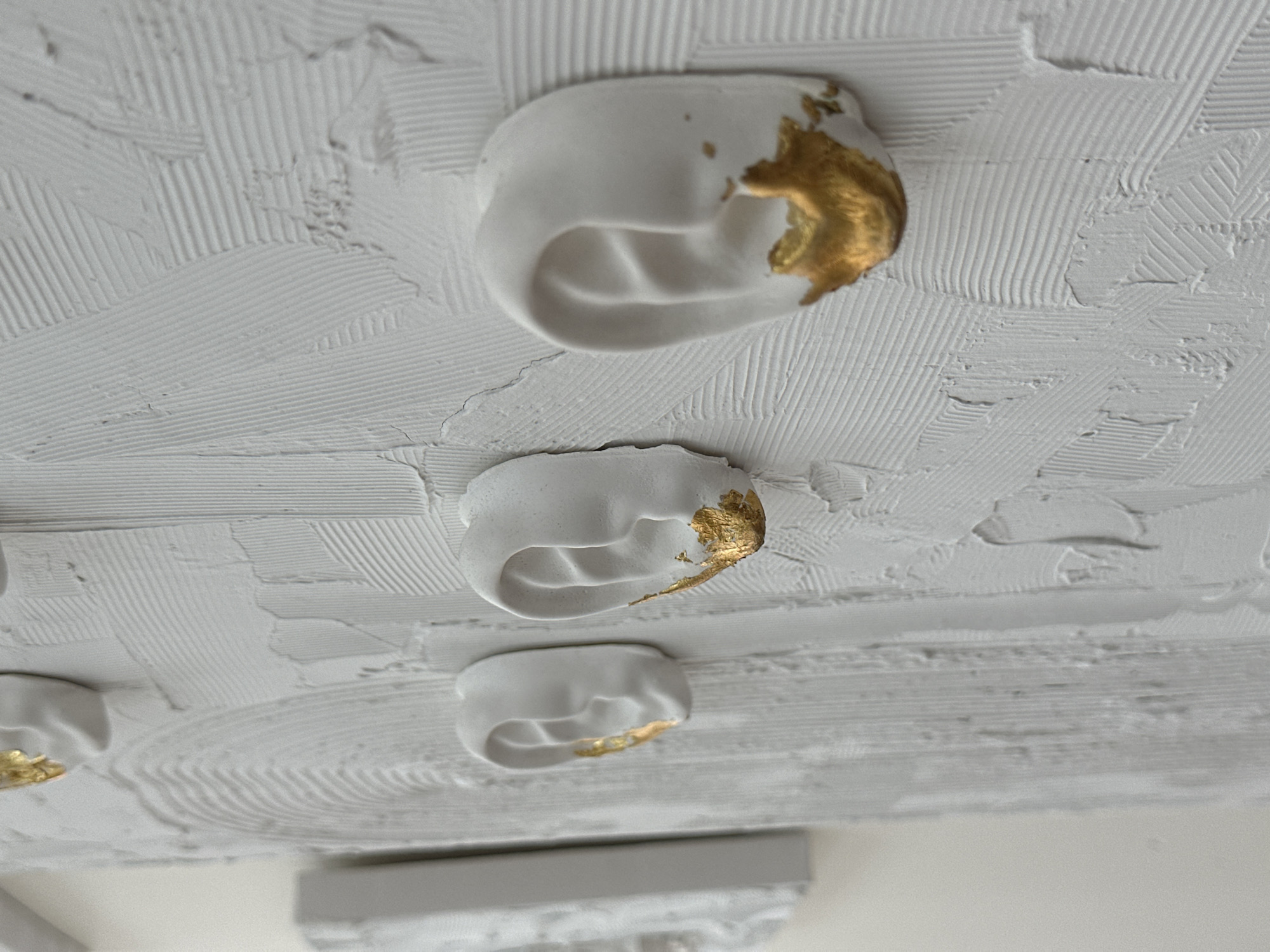 The image shows a close-up of a textured white surface, likely a ceiling or wall. Three rounded, white, semi-oval protrusions with gold leaf accents are embedded within the texture. The surface texture consists of linear, ridged patterns resembling trowel marks. Based on the provided information, it is impossible to confirm the image's location in Sochi, Russia.