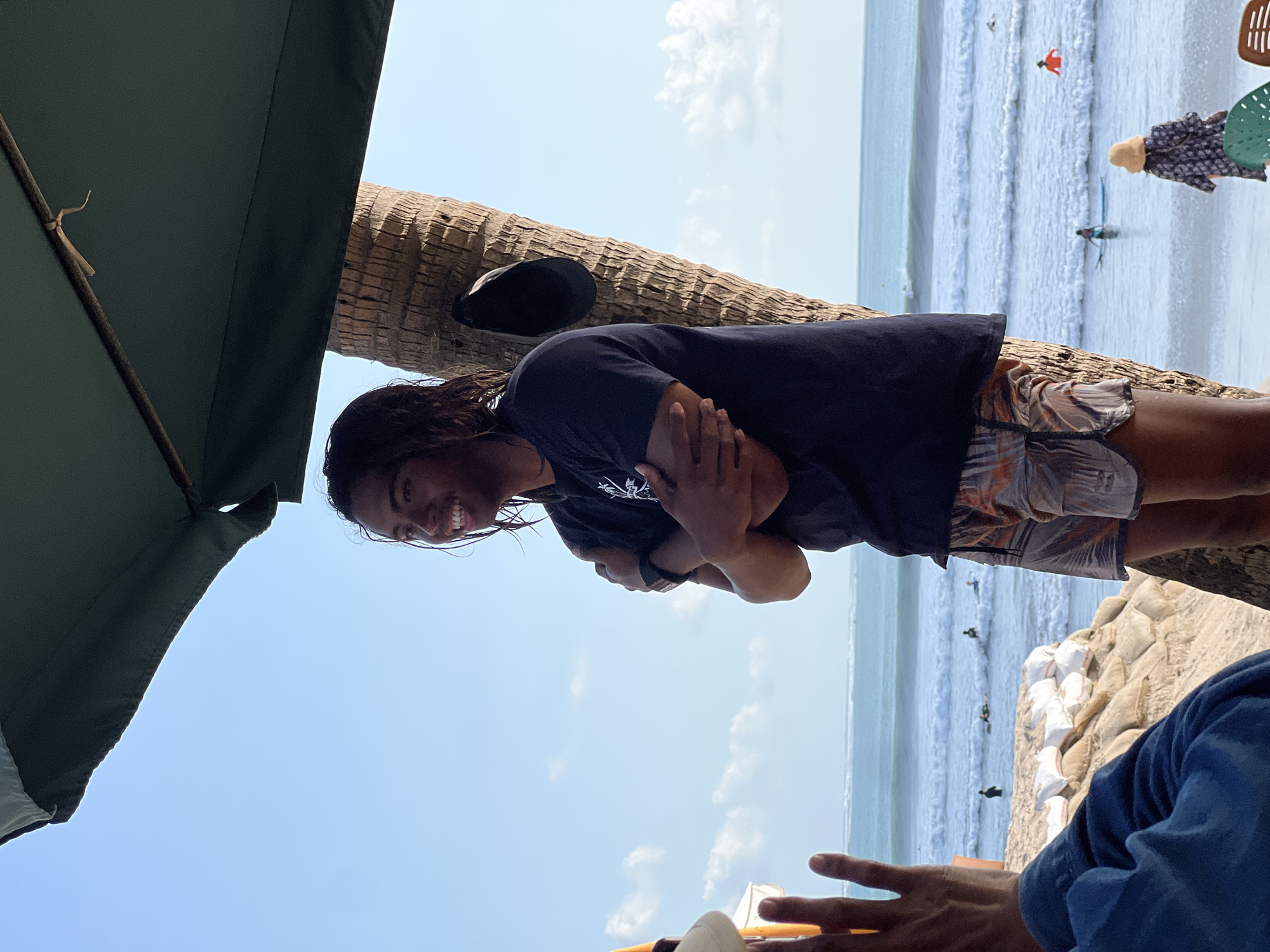 A young man with long, dark, wet hair stands on a beach in Kuta, Indonesia, wearing a dark t-shirt and patterned shorts. He has his arms crossed and is smiling. He is positioned next to a palm tree, on which a dark baseball cap is hanging. In the background, the ocean is visible with several surfers in the water. Sandbags are piled up on the beach in front of the water. A large, dark green umbrella provides shade overhead. A person's hand, with fingers extended, is in the foreground on the left side of the image. Another person wearing a blue shirt is partially visible in the lower left corner. A woman wearing a patterned dress and a straw hat is visible on the beach on the right side of the image, facing away from the camera.