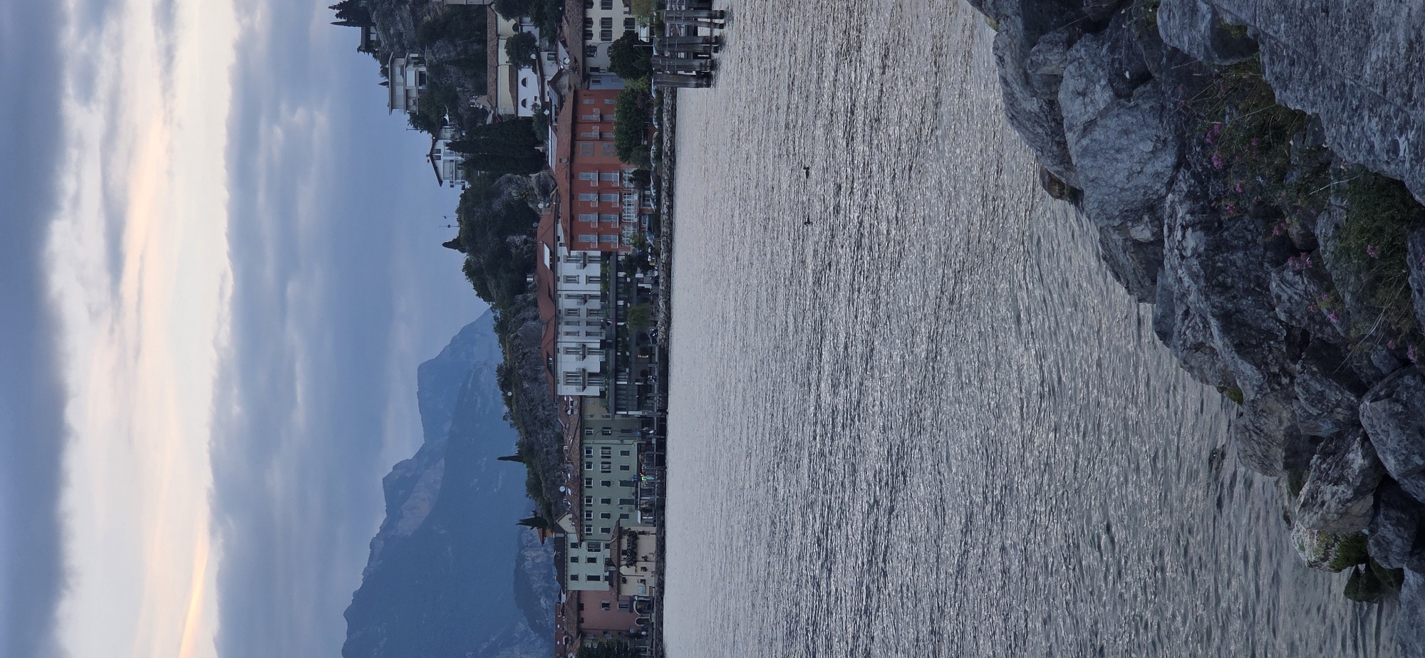 This image depicts a waterfront view of the town of Torbole, Italy, located on the shores of Lake Garda. The foreground features a rocky shoreline with scattered greenery and small pink flowers. Beyond the shore lies the calm, rippling water of the lake, reflecting the overcast sky. In the middle ground, a line of multi-story buildings with colorful facades—ranging from pale green and yellow to orange and white—forms the town's waterfront. These buildings appear to be a mix of residential and commercial structures, with visible windows and balconies. Further in the background, steep, rugged mountains rise, their peaks softened by the hazy, twilight atmosphere and overlaid with layers of dark, diffused clouds. A few scattered trees are visible on the slopes. The overall scene is characterized by muted colors and a serene, slightly melancholic ambiance, suggesting it was captured during dusk or early morning. There are no people visible in the image. Two ducks are visible swimming in the lake.