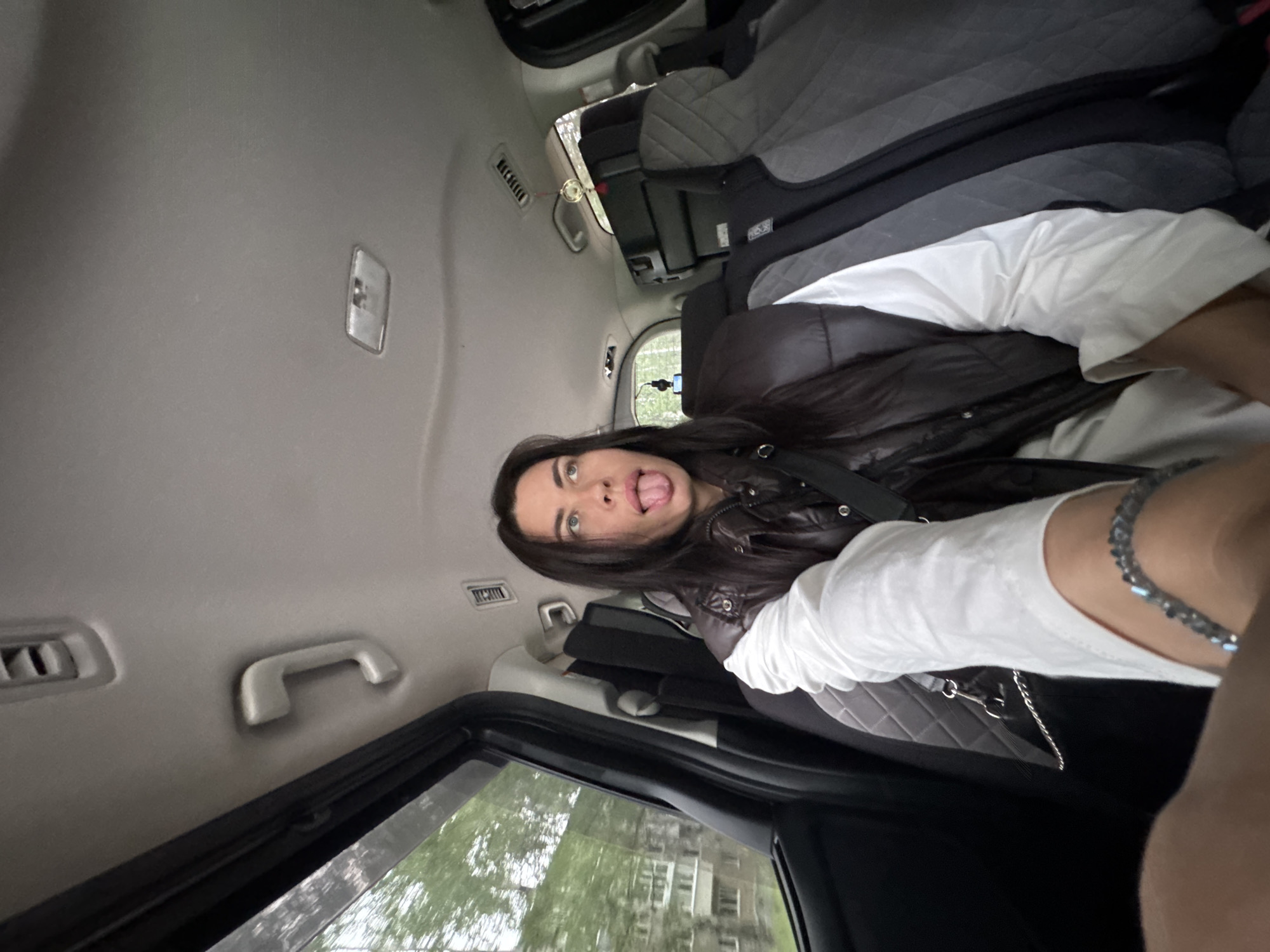 A woman is sitting in the back seat of a car, sticking her tongue out at the camera. She has long, dark hair and is wearing a white shirt and a dark vest. The car appears to be moving, as evidenced by the blurry trees and buildings visible through the window on the left side of the image. The interior of the car is visible, including the seats, headrests, and ceiling with overhead lights and handles. Stacked luggage is visible in the back of the car behind the woman. The scene appears to be in a residential area, possibly in the city of Chaikovsky, Russia, given the architecture and vegetation.