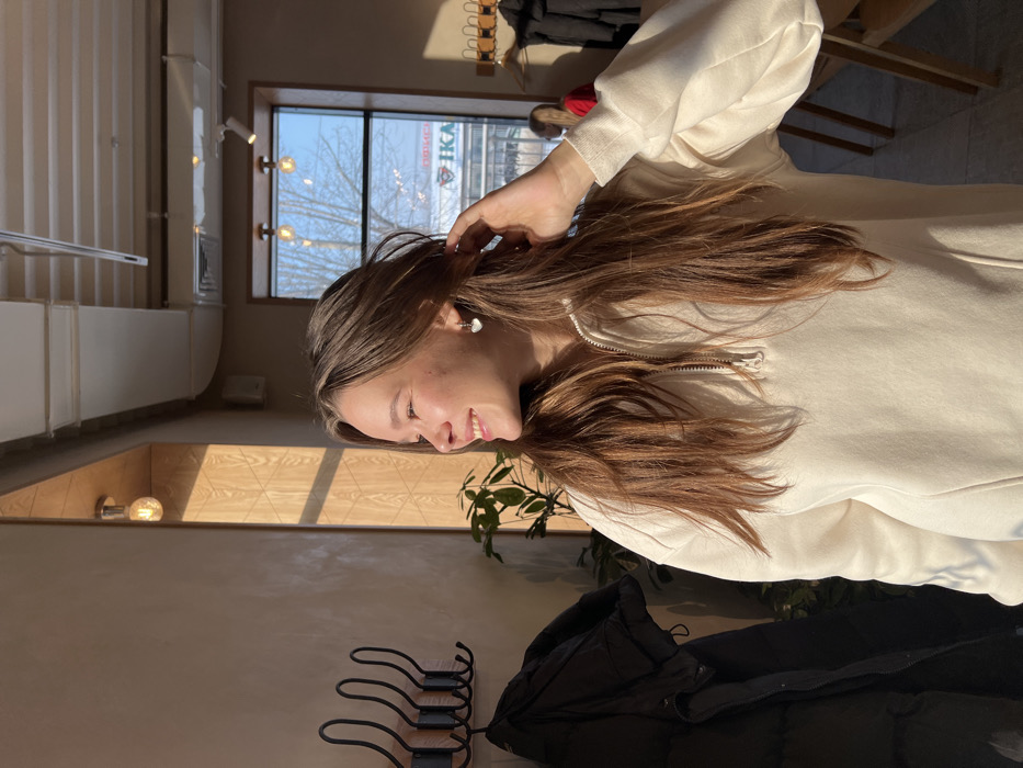 A young woman with long brown hair smiles, her hand near her ear, wearing a cream-colored zip-up sweater. A window behind her reveals a building with signage. The interior includes a wooden wall with a light fixture and a coat rack holding a black jacket. Greenery is visible near the woman. The scene is well-lit with natural sunlight.