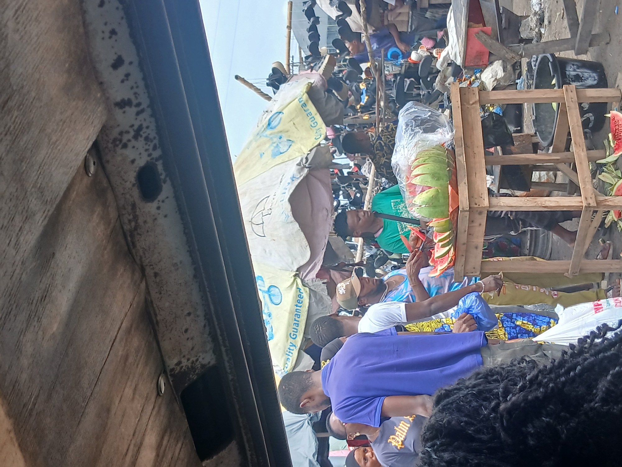 This is a media file depicting a bustling outdoor market scene in Agani, Nigeria. The image is oriented vertically, and the foreground is dominated by the side of a wooden structure, possibly a truck bed, with visible wear and tear.
The main subjects are a group of people, appearing to be vendors and customers. One man in a bright green shirt is prominent, holding a long, dark object that resembles a stick or a tool. He is surrounded by various goods, including sliced watermelons displayed in a plastic bag and on a wooden structure. Other individuals are seen interacting within the crowded market. Some are wearing casual clothing, including a blue t-shirt and a patterned fabric. The background reveals more stalls, some covered with tarps, displaying shoes and other items hanging from racks. Large black containers are also visible.
The activity taking place is a commercial exchange, typical of a marketplace. People are gathered, likely buying and selling goods. The presence of sliced watermelons and shoes suggests a food vendor and a shoe vendor.
Notable details include the bright sunlight, indicating it is daytime. The sky is visible and appears clear. The setting is clearly an open-air market, characterized by makeshift stalls and a dense gathering of people and goods. There is a sense of busyness and activity.
Visible text includes the words "City Guarantees" on a yellow tarp, and partially visible text "PALM" on a t-shirt in the lower part of the image. The overall impression is a vibrant, energetic market day.