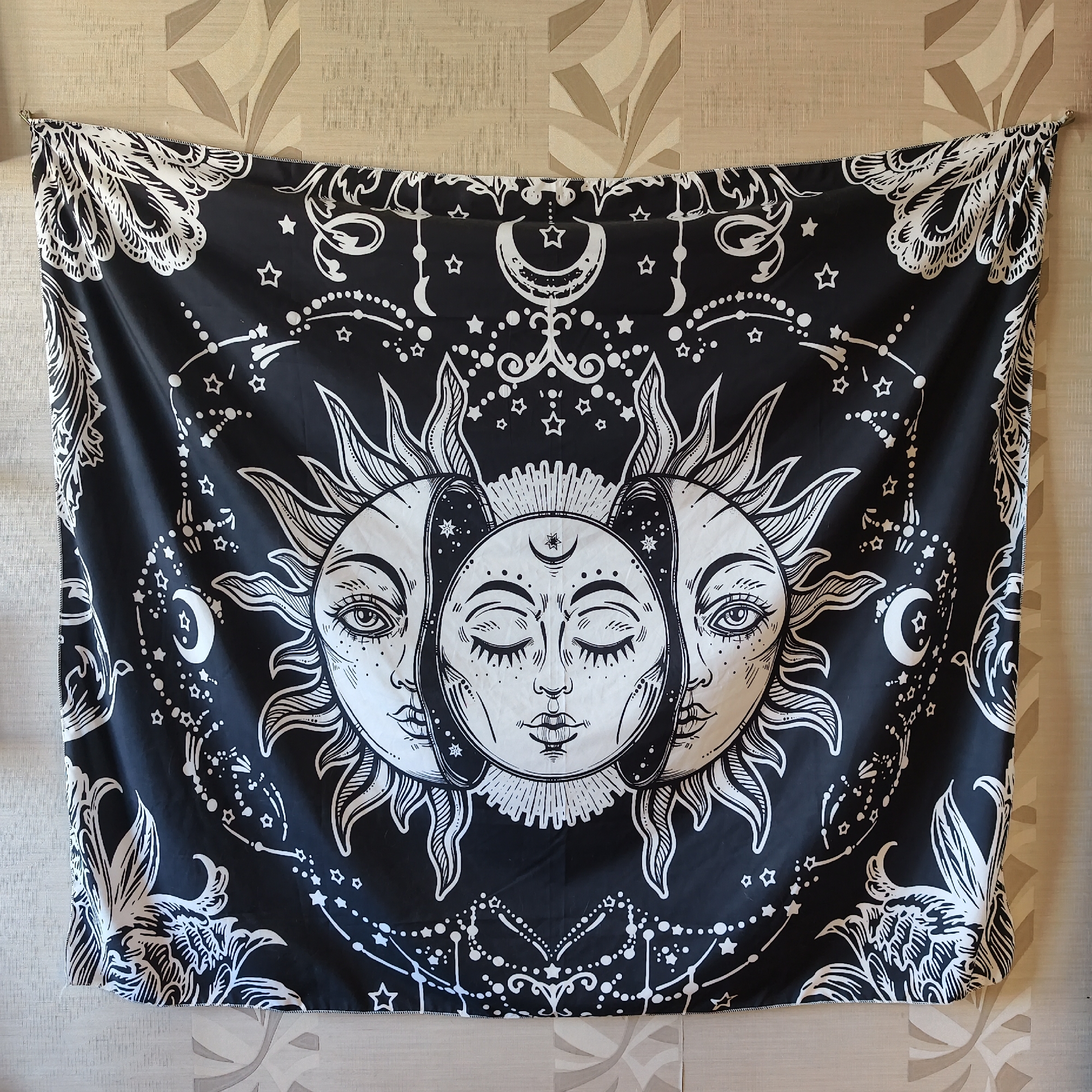 The image shows a black and white tapestry hanging on a wall with a textured wallpaper. The tapestry features a celestial design with three faces in the center, resembling phases of the moon or sun. The central face is closed-eyed and serene, while the two side faces are open-eyed and looking outward. These faces are surrounded by stylized sun rays and various celestial elements like stars, crescent moons, and constellations. The borders of the tapestry are adorned with intricate patterns of leaves and feathers. The tapestry is secured at the top with small metal clips, suggesting it is displayed as wall art. The lighting suggests an indoor setting. No visible text is present on the tapestry or in the environment. The wallpaper has a beige and brown abstract pattern.