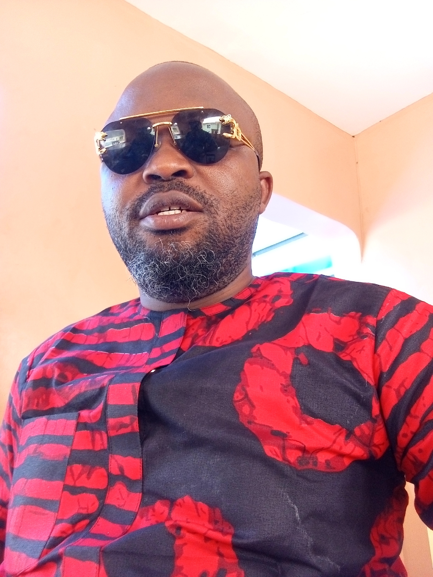 A medium shot captures a bald, brown-skinned man with a salt-and-pepper beard wearing gold-rimmed sunglasses. He is dressed in a red and black patterned shirt. The background is a light peach wall. The location is presumably indoors in the city of Ankpa, Nigeria, although this cannot be verified from the image. There is no visible evidence to confirm the location.