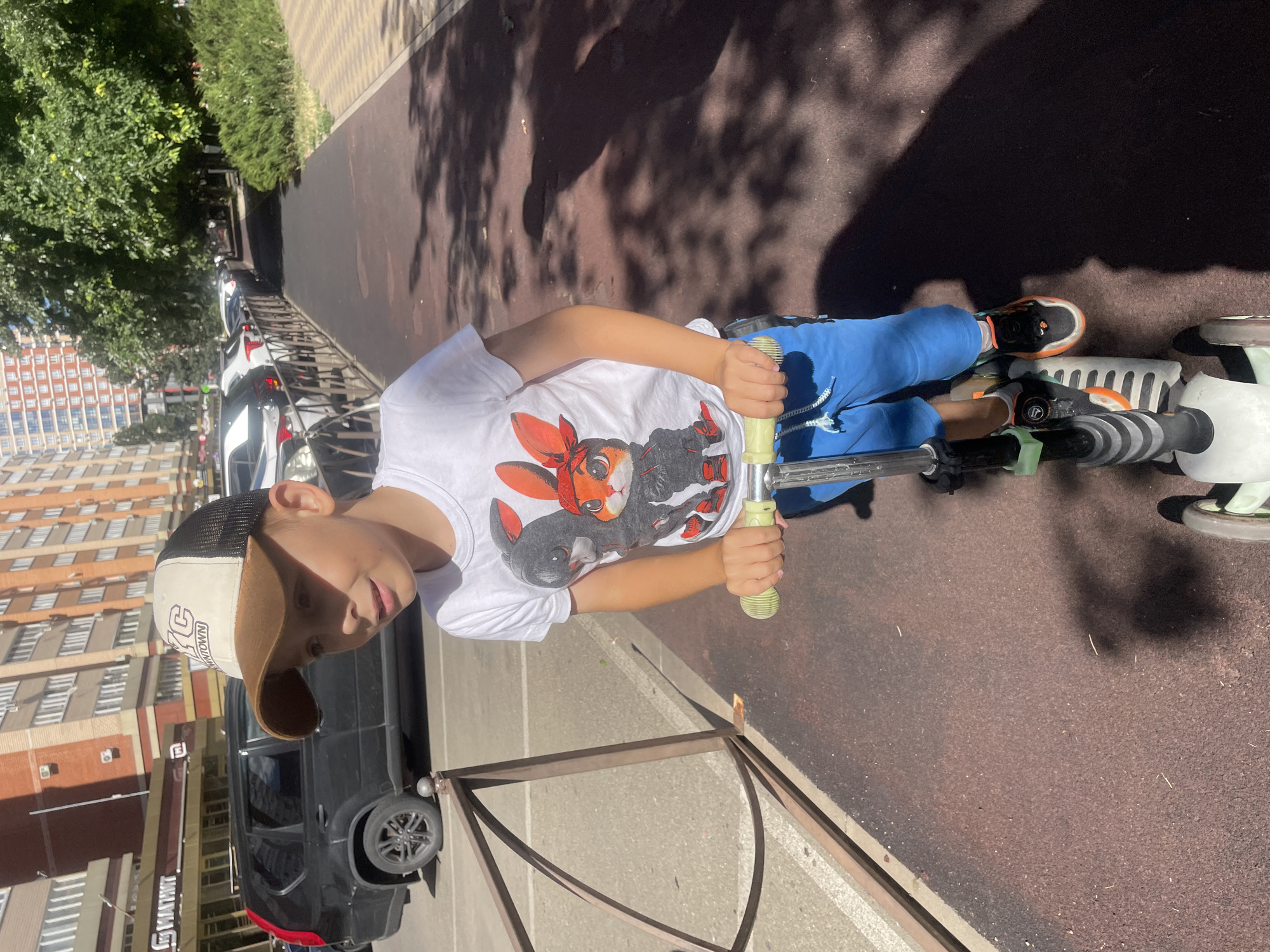 A young boy, approximately 6-8 years old, wearing a white t-shirt with cartoon rabbits, blue shorts, and black and orange sneakers, is riding a three-wheeled scooter on a paved path. He has a brown and beige baseball cap on his head. The background shows a street with parked cars, including a black SUV, and multi-story buildings. Trees and shadows are also visible. The image appears to be taken in Krasnodar, Russia, based on the architectural style of the buildings and the general environment.