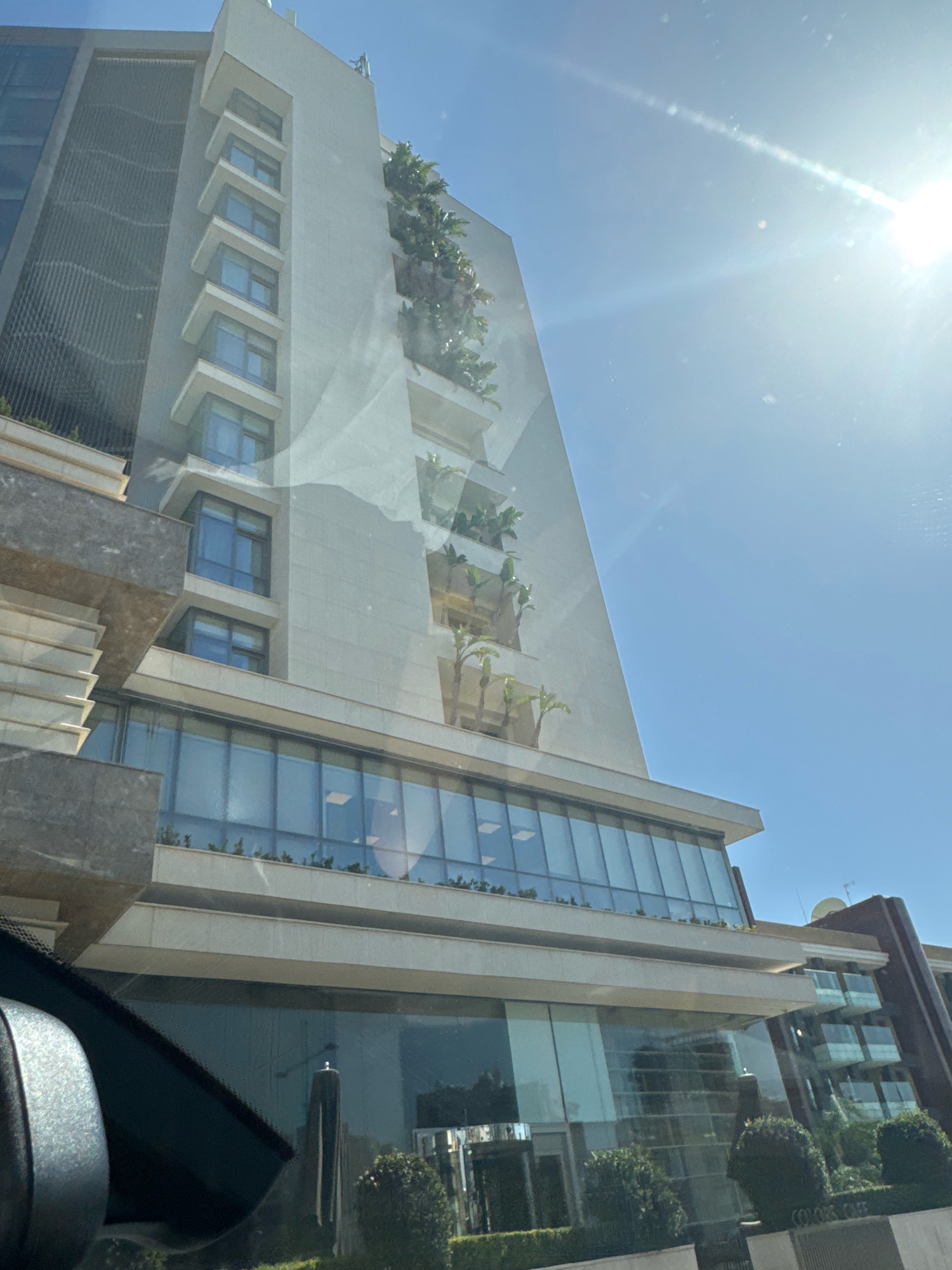 The image shows a modern building with a light-colored facade and many windows, situated in Agios Tychona Community, Cyprus. The building has several tiers with balconies and is adorned with greenery, suggesting a design incorporating nature. A bright sun is visible in the upper right corner, casting a lens flare across the sky. The lower part of the image shows a street-level view with glass doors and surrounding greenery. The reflection in the glass indicates the presence of trees and possibly other buildings. The overall scene depicts an urban environment with a contemporary architectural style.