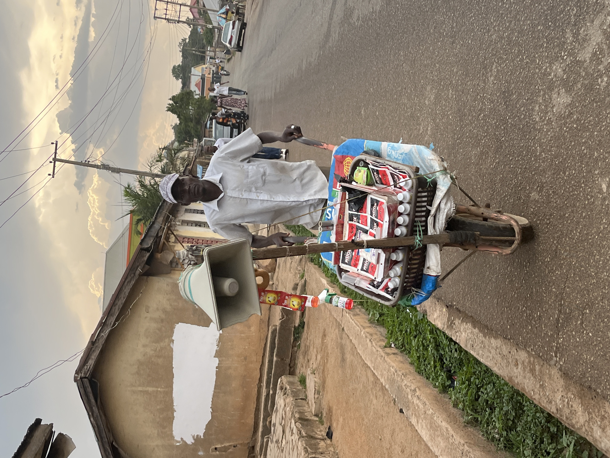A man in a white shirt and cap stands on a street in Jos, Nigeria, with a handcart loaded with goods for sale. He is facing the camera and holding the cart's handlebar. A megaphone is mounted on the cart's side. The cart contains packets of "Pestox" and white plastic containers. The street is paved, and there are buildings and power lines visible in the background. Other people and vehicles are visible in the distance on the street. The sky is overcast.