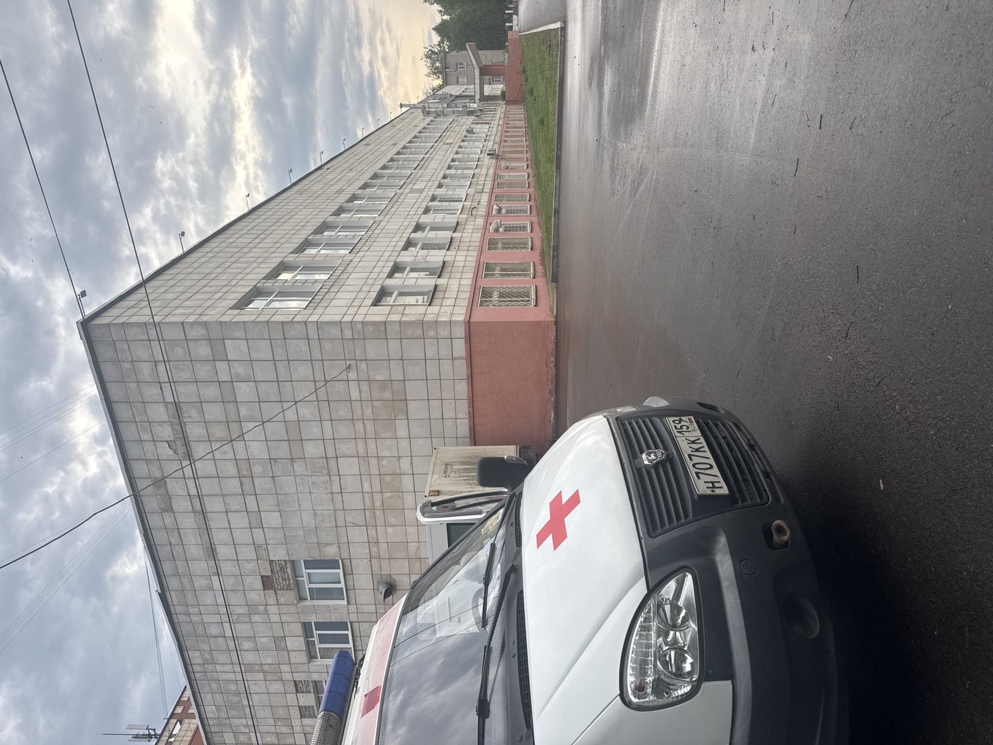 A low-angle, medium shot depicts a white ambulance parked on a wet, dark asphalt road in front of a large, rectangular building. The building, possibly a hospital, features a tile exterior with multiple rows of windows. The lower section is painted in a pinkish-red hue. A strip of grass runs along the building's base. Overcast skies are visible above the building. The ambulance, marked with a red cross on its hood, has a visible license plate "H707KK159", indicating the vehicle is registered in the Perm Krai region of Russia, where Chaikovsky is located. Based on the ambulance and the large building, the location is likely near a medical facility in Chaikovsky, Russia. No people are directly visible in the frame.