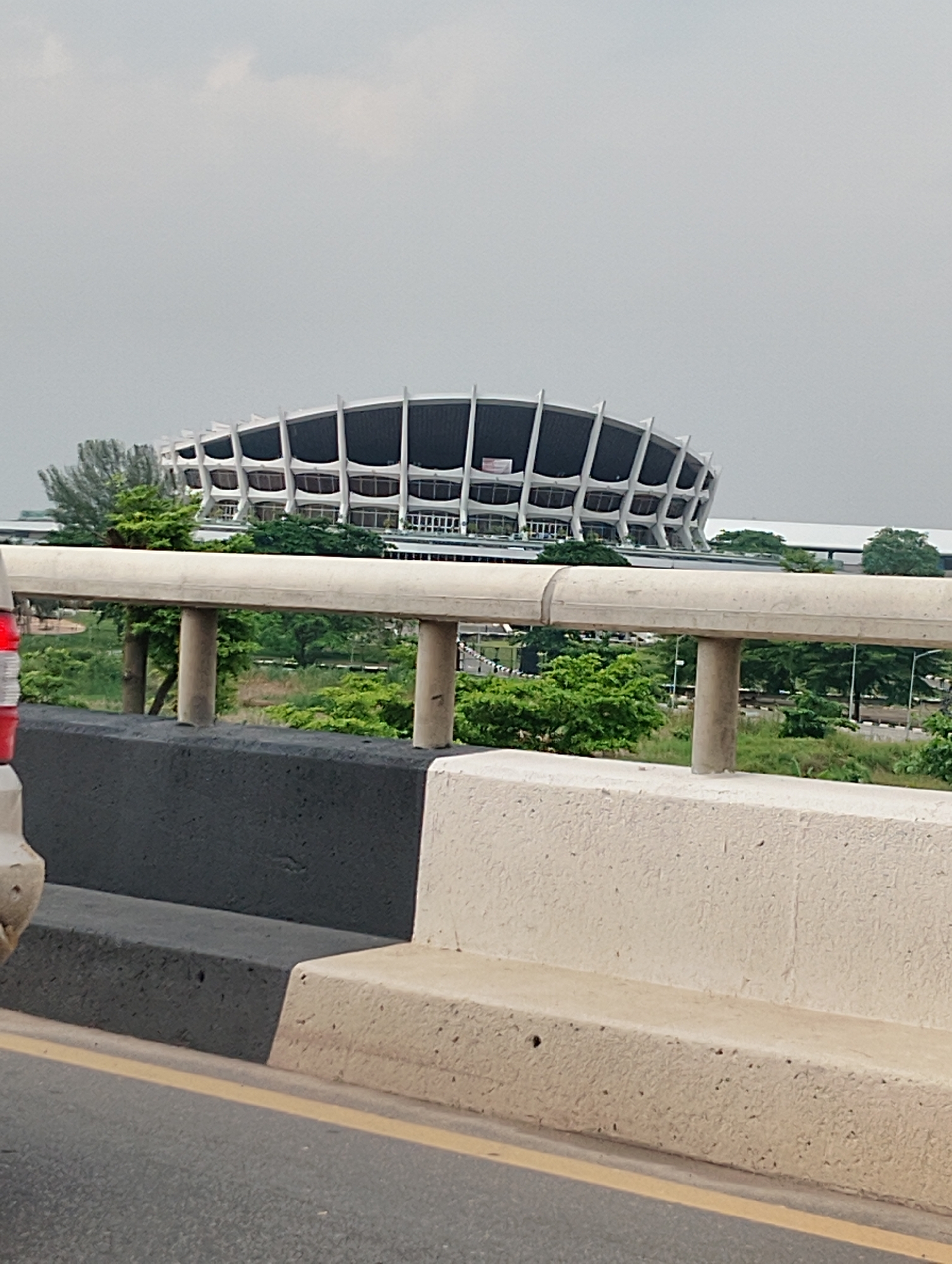 The image captures a view from a moving vehicle on a highway, looking towards the National Arts Theatre in Alagbede, Nigeria. In the foreground, a concrete barrier with a dark gray lower section and a lighter beige upper section separates the road from the landscape beyond. A yellow line runs along the edge of the road. Behind the barrier, lush green trees and vegetation are visible. In the mid-ground, the iconic National Arts Theatre stands out with its distinctive curvilinear roof supported by a series of white pillars. The building has multiple levels with rows of windows. The sky is overcast, with a light gray haze. There are no people visible in the image.