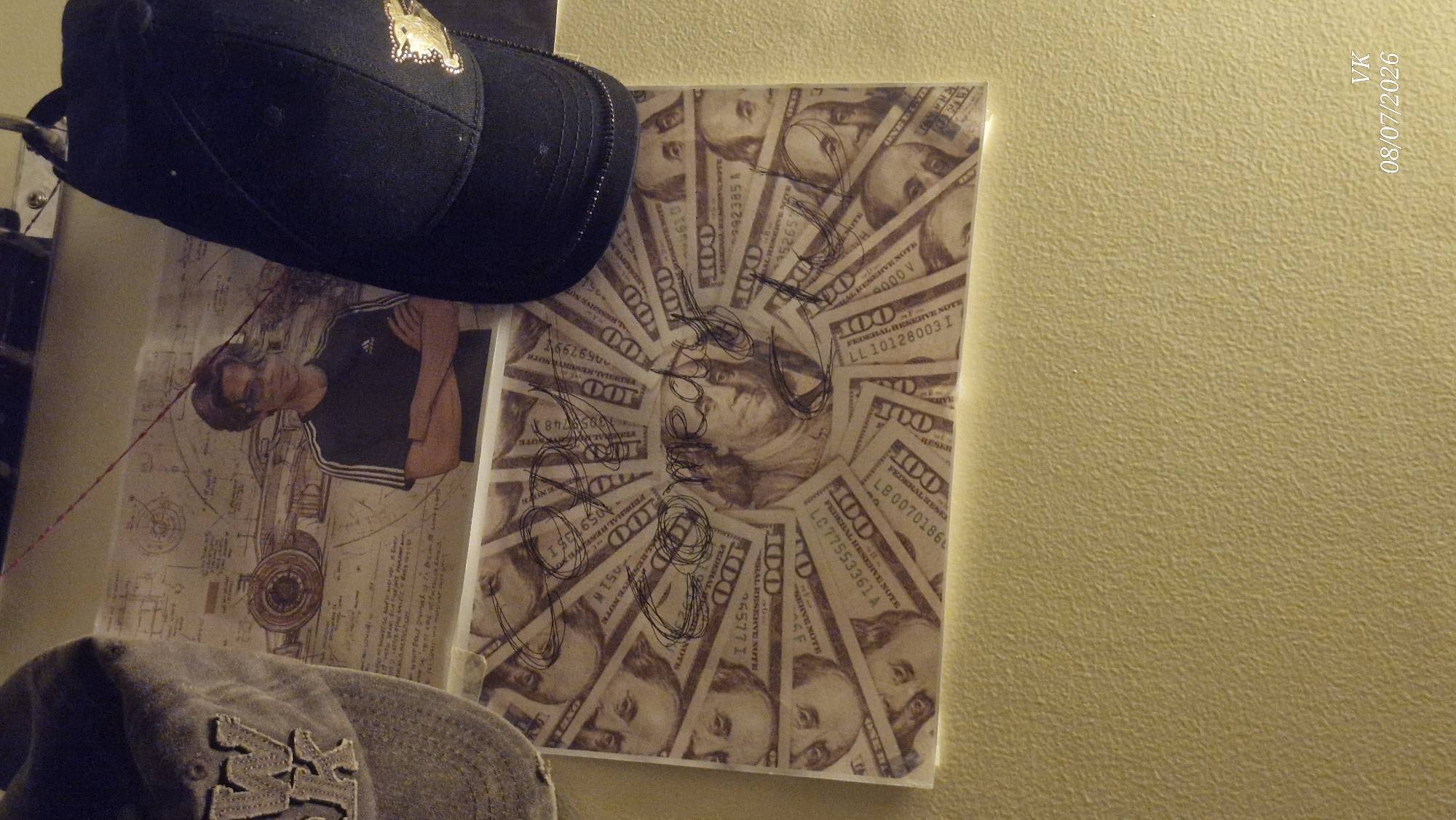 The image shows a close-up of a wall with a textured yellow surface. In the upper portion of the image, there are three objects: a grey baseball cap on the left, a poster or print in the middle featuring a blueprint of a car and a young man wearing a black Adidas t-shirt with his arms crossed, and a black baseball cap with a gold emblem on the right. Below the poster is a rectangular print of US $100 bills arranged in a circular pattern, with some scribbled text in black marker over it. The text appears to say "Comed y Comed y" or something similar. In the bottom right corner of the image, the text "VK 08/07/2026" is visible. This image is not located in Vrindavan, India.