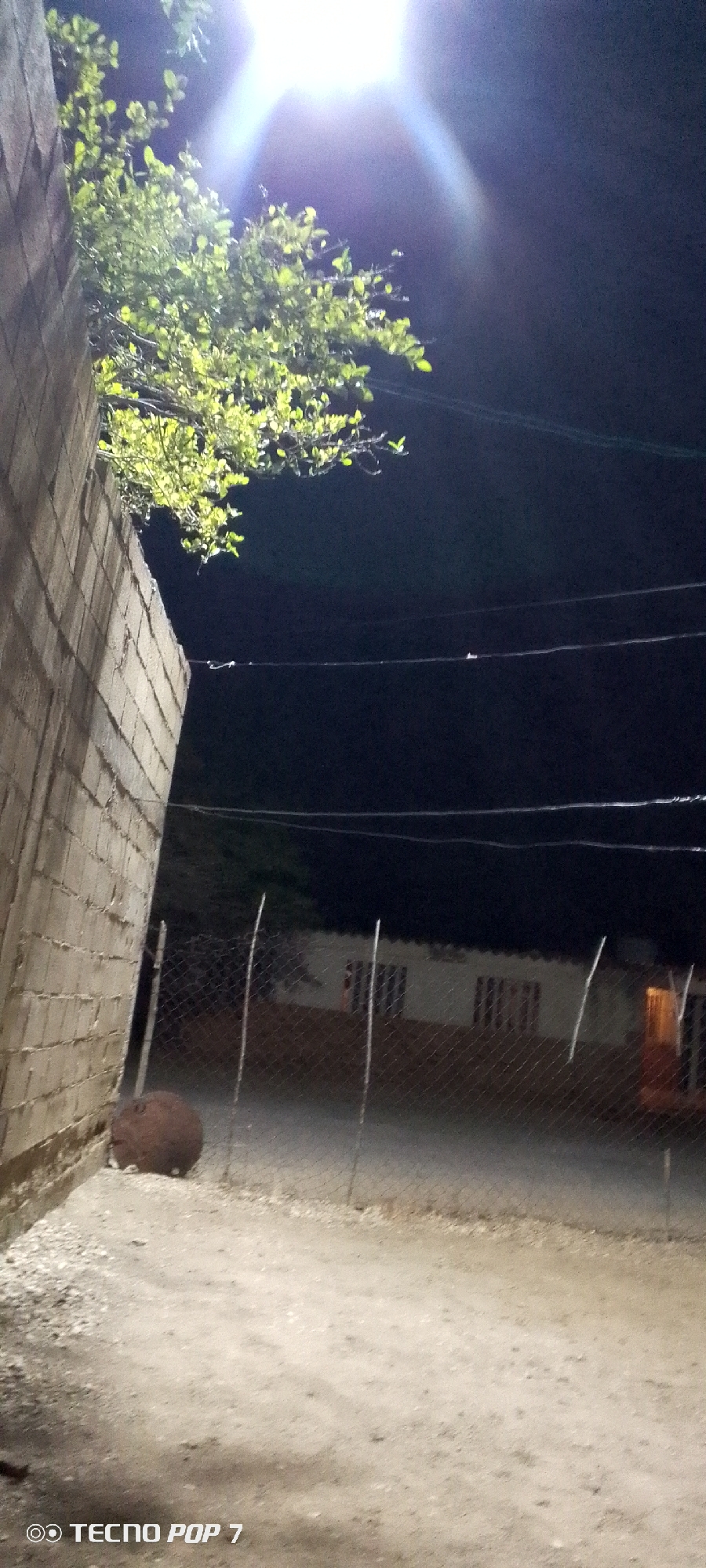 The image presents a nighttime scene in what appears to be a residential area of Altagracia, Venezuela. A building is visible, constructed from cinder blocks or a similar material. To the left of the building, a large, brown, round object is situated, possibly a boulder. In the background, a white structure with windows is partially obscured by a wire fence. Power lines span the upper portion of the scene against the dark sky. A bright light source illuminates the scene. Vegetation is also present, partially obscuring the background. The ground appears to be dirt or gravel. The "Tecno Pop 7" watermark is visible in the bottom left corner of the image. No people are visible.