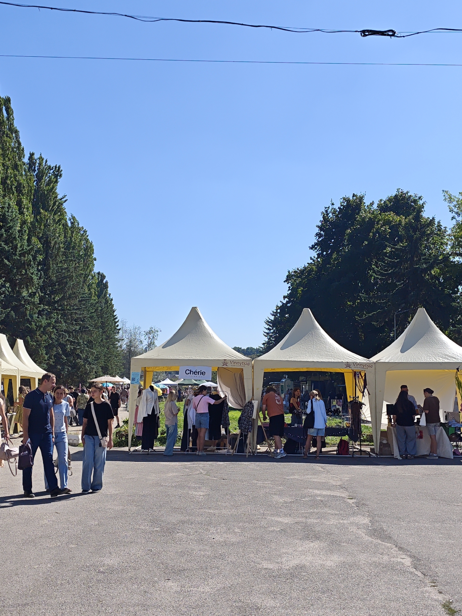 The image shows an outdoor market in Vinnytsia, Ukraine, under a clear blue sky. Several white, conical tents are set up, serving as stalls for vendors. In the foreground, a paved walkway is visible, with a few people walking. In the mid-ground, a row of tents displays clothing on racks and tables, with shoppers browsing. People are seen interacting with vendors and examining merchandise. Tall, green trees line the left side of the scene, and more trees are visible in the background behind the tents. The overall impression is of a lively community event.