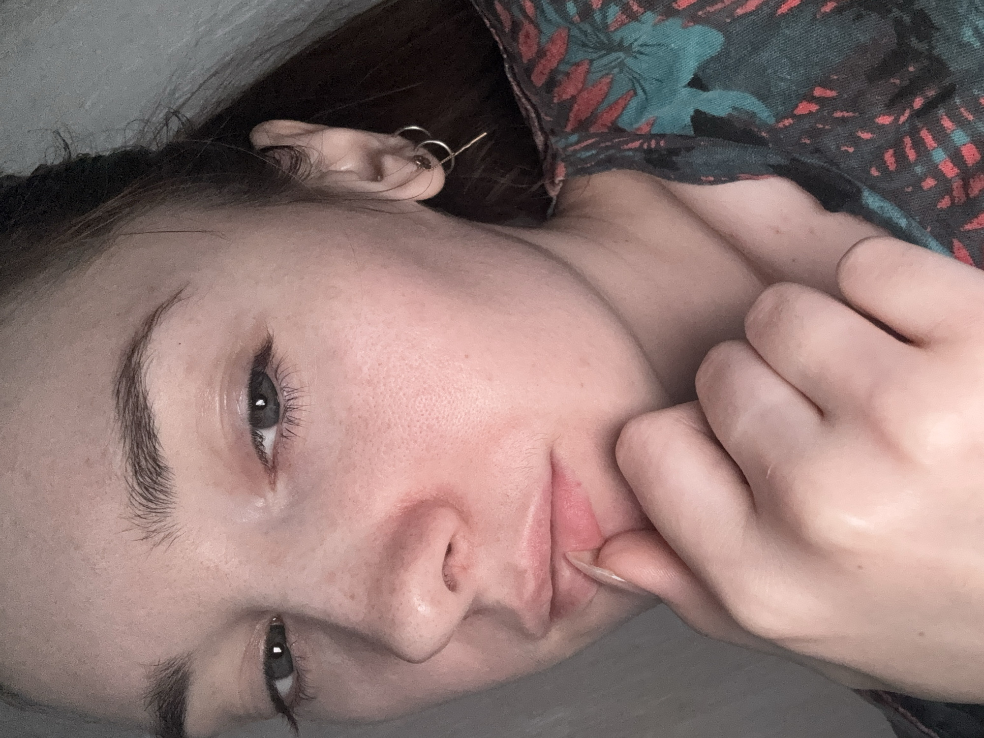 A young Caucasian woman with light skin, freckles, and dark hair is captured in a close-up, self-taken portrait. Her eyes are looking towards the camera, and her right hand is resting on her chin, with her index finger touching her lips. She is wearing a multi-colored, patterned top. The lighting suggests an indoor setting with a neutral background. There is no indication of the city of Opochka, Russia, in the image.