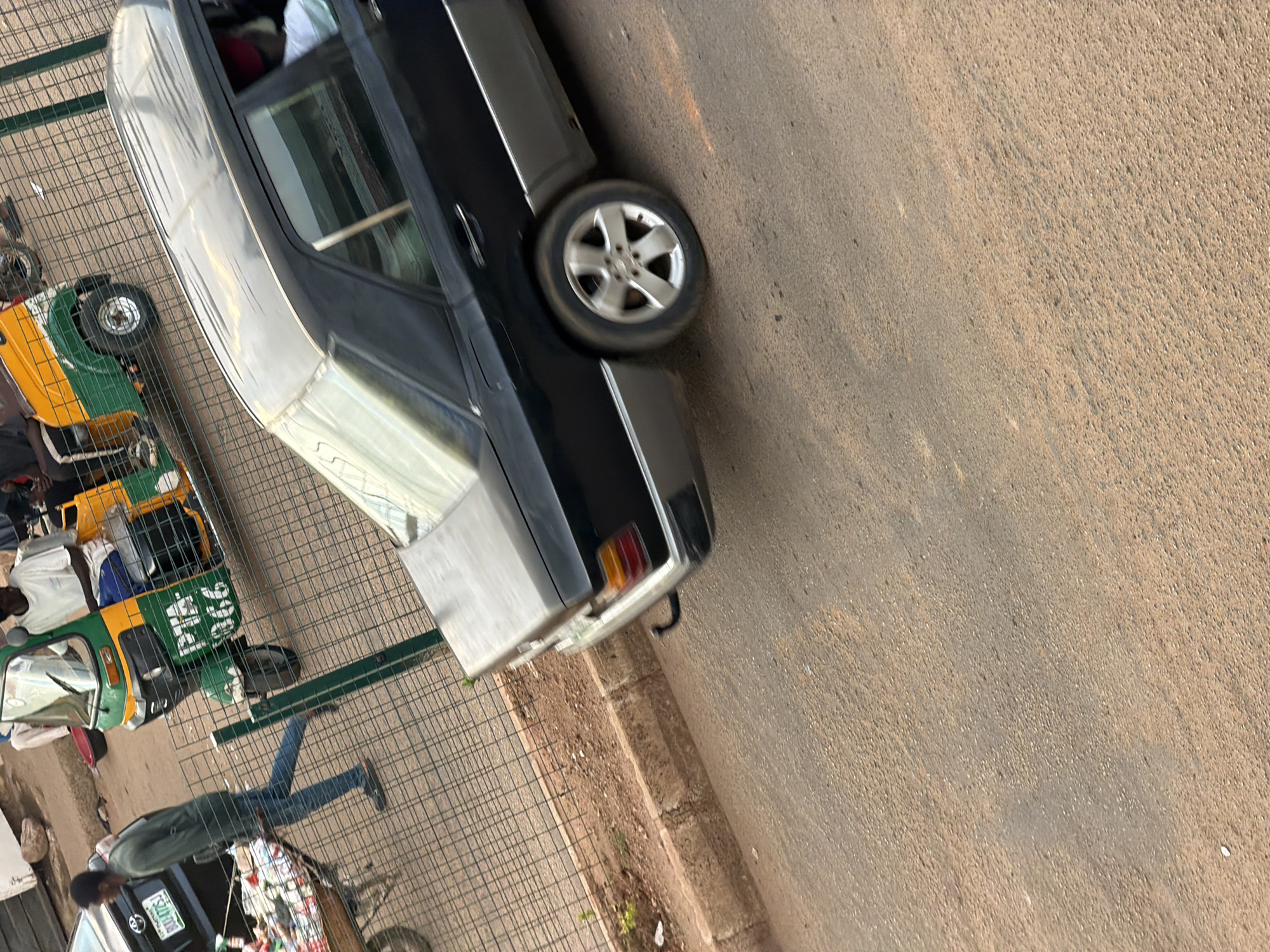 In the city of Bukuru, Nigeria, an outdoor scene unfolds on a dusty street. A black Mercedes-Benz sedan with a silver lower body panel is parked on the right side of the frame. Its rear wheel, a five-spoke alloy, is visible and slightly blurred, indicating movement or a shallow depth of field. The car has a black roof and tinted windows.
To the left of the sedan, a man in a dark green long-sleeved shirt and blue jeans pushes a cart loaded with goods. The cart, a makeshift arrangement with a wooden base and metal frame, is filled with various items, including what appear to be food products wrapped in plastic. A black utility vehicle with the license plate "BOD-7053" is partially visible behind the man.
In the background, a vibrant green and yellow auto-rickshaw, also known as a Keke Napep, is parked. The auto-rickshaw has "PIA" and "366" written on its side in white lettering. Two individuals are seated within the auto-rickshaw. One person, wearing a white t-shirt, is visible in the driver's seat. Another person, partially obscured, is seated behind them.
The ground is a mix of paved and unpaved surfaces, with a visible curb. A wire fence runs along the left side of the frame, separating the street from a more sandy and uneven area where sparse vegetation grows. The lighting suggests it is daytime, possibly late afternoon, with shadows cast on the ground. The overall impression is a snapshot of daily life and transportation in a Nigerian urban environment.