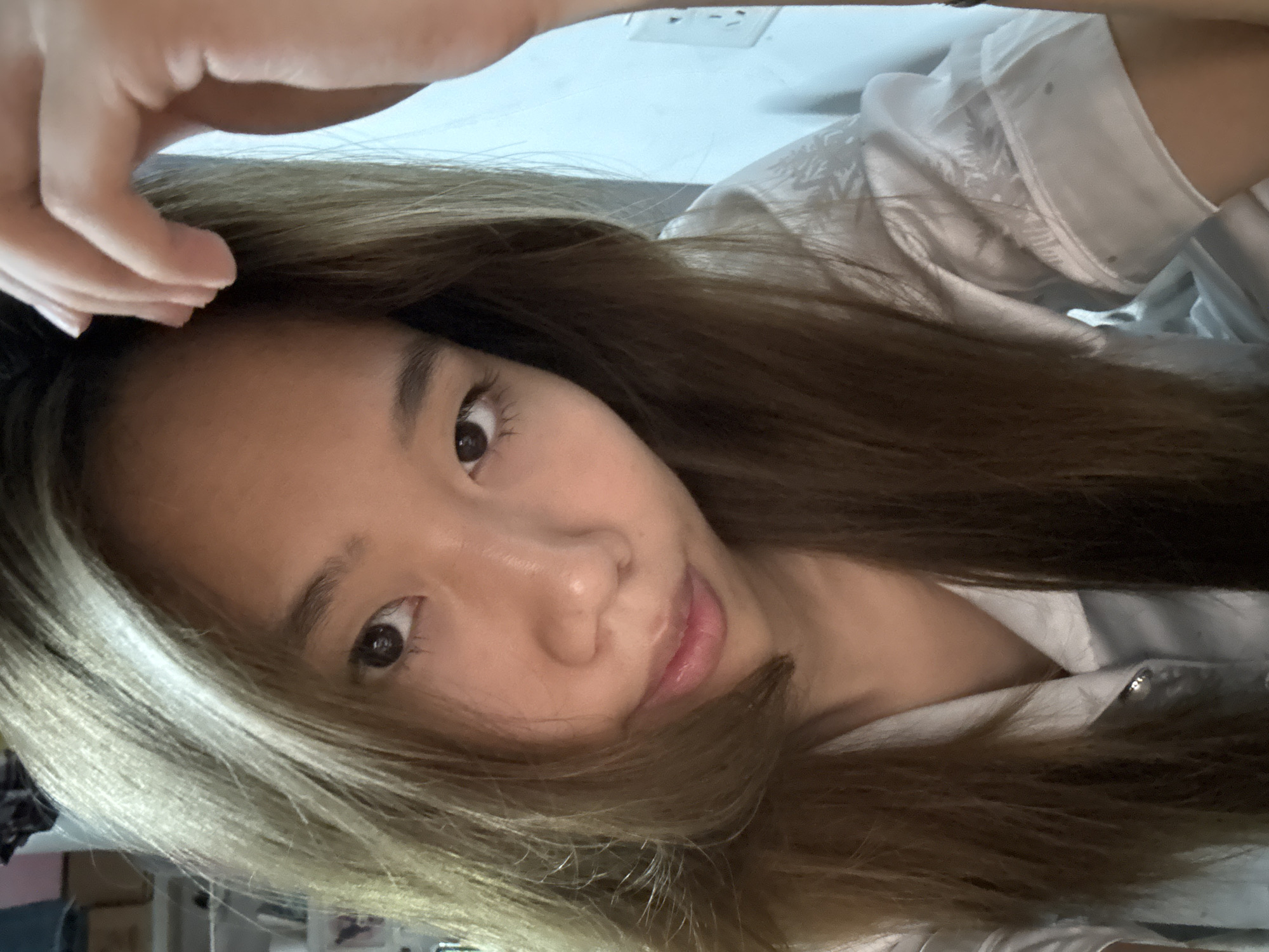 A young woman with light brown hair is posing for a selfie. Her head is tilted slightly, and she is looking directly at the camera with a small smile. Her left hand is raised towards her forehead. She is wearing a white shirt with a button visible at the neckline. In the background, there's a light blue wall, and a partially visible electrical outlet. The location in the city of Nan'an District, China, is not discernible from the image.