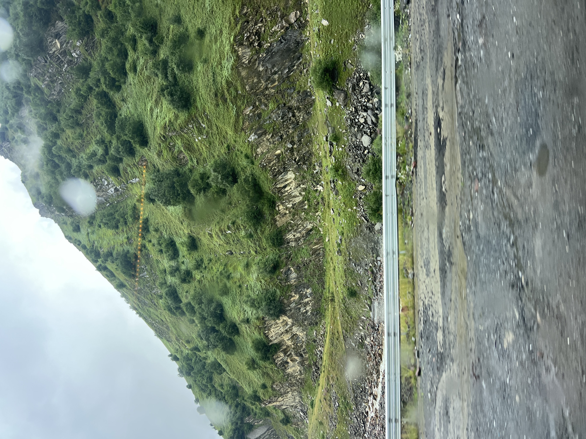 The image depicts a mountainous landscape in Almasiani, Georgia, under a cloudy sky with visible raindrops on the lens. A steep, green hillside covered in trees and rocks dominates the upper portion of the frame. A silver guardrail runs horizontally across the middle of the image, separating the hillside from a wet, rocky terrain and a road in the foreground. The road surface is dark and appears to have puddles of water. The overall scene suggests a remote, natural environment, possibly along a road leading through the mountains.