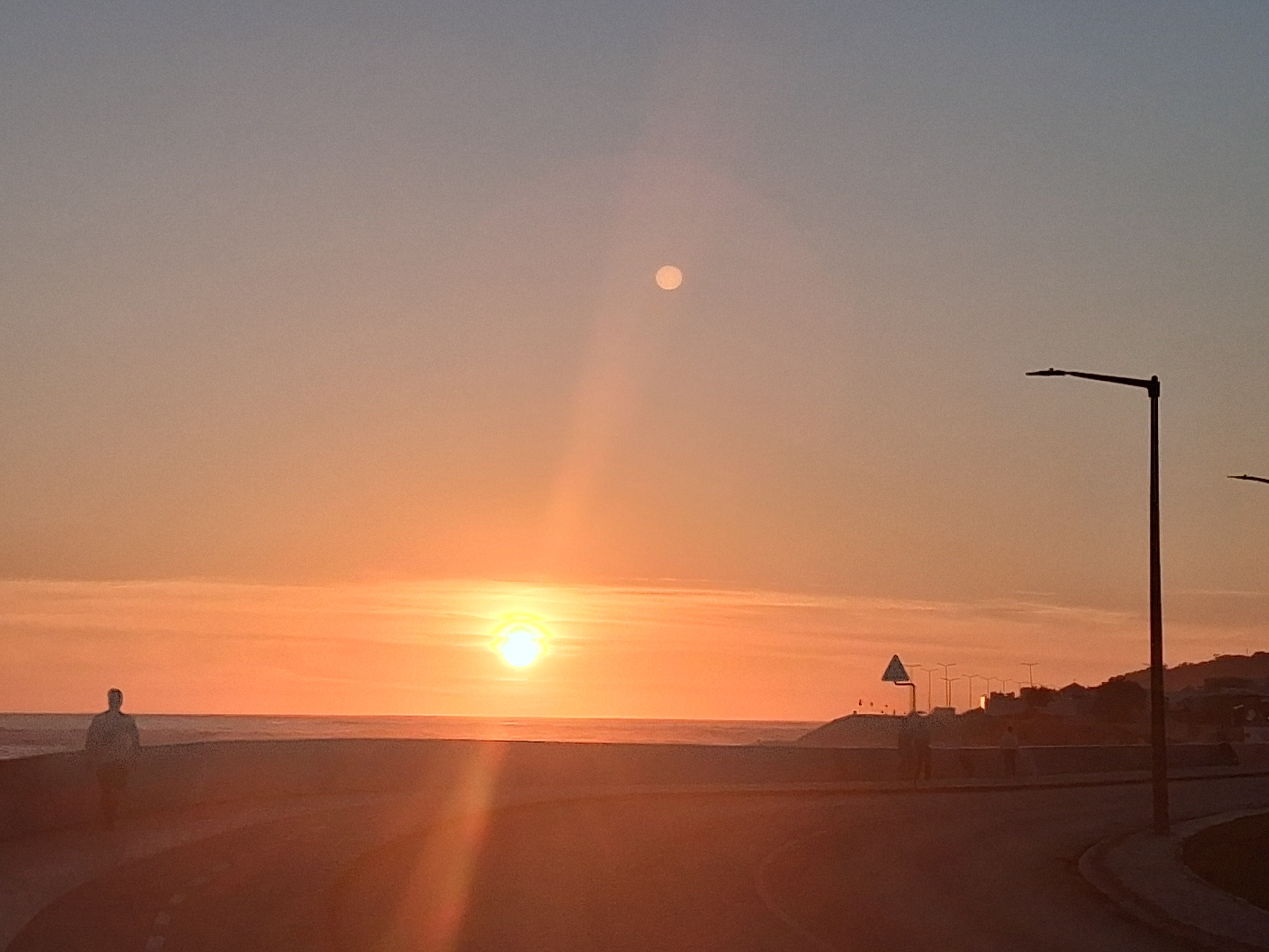 The image captures a sunset scene in Figueira da Foz, Portugal. The sun is setting over the Atlantic Ocean, creating a strong glare and a vertical beam of light on the road. Above the sun is a small, pale circular object, possibly the moon or a lens flare. A person is walking along the beach on the left side of the image. On the right side, there is a street with street lamps. A series of buildings and other street lamps can be seen in the distance on a slight hill. The foreground consists of the road surface. The overall color palette is dominated by orange and yellow hues from the sunset.