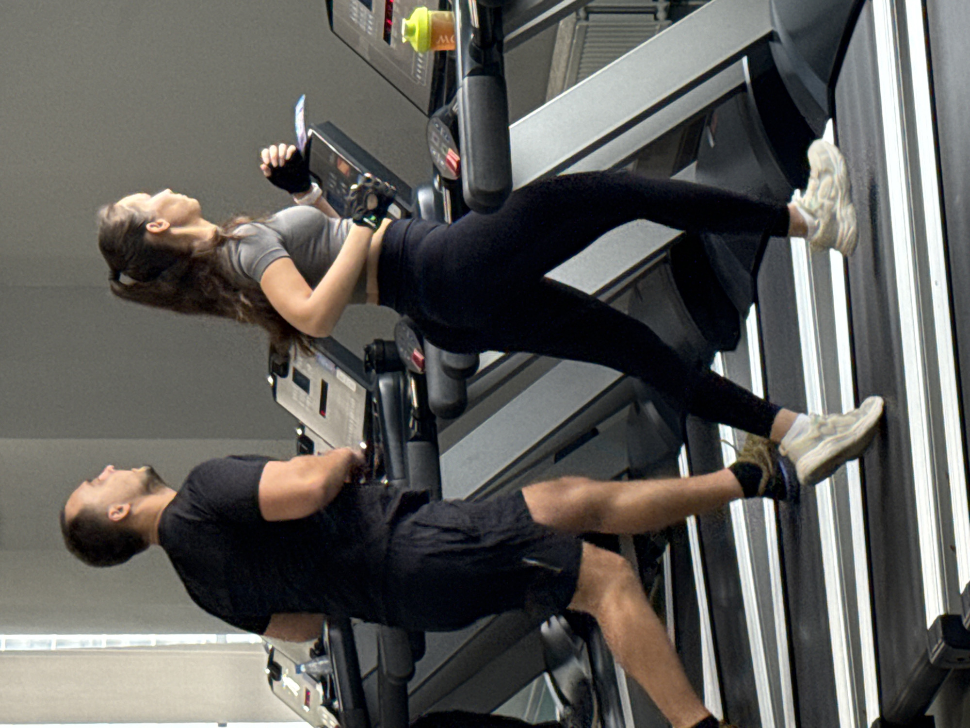 This image captures two individuals, a woman and a man, exercising on separate treadmills in an indoor gym in Yekaterinburg, Russia. The photograph is oriented vertically, with the subjects appearing to stand upright relative to the frame.
In the upper half of the image, a woman with long, dark brown hair is visible. She is dressed in a light grey short-sleeved crop top, black leggings, white sneakers, and black fingerless gloves. She holds a smartphone in her left hand and appears to be looking at its screen. A bright yellow water bottle with an orange cap rests on the console of her treadmill.
Below her, a man is seen on an adjacent treadmill. He has short dark hair and a beard, wearing a black t-shirt, black shorts, and dark sneakers with light-colored soles. He is looking upwards or straight ahead.
Both treadmills feature digital display consoles, though no specific text is clearly legible on them. The background is a plain, light grey wall or ceiling, characteristic of an indoor fitness facility. The lighting is artificial, common in gyms. The overall scene suggests a routine workout session, with both individuals focused on their activity or personal devices. The exact time of day is not discernible due to the indoor setting.