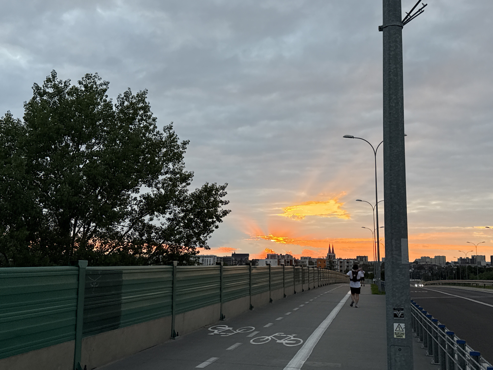 The image shows a pedestrian and cyclist path on a bridge over a highway in Białystok, Poland, at sunset. A person is running on the path, facing away from the camera. The sky is cloudy with vibrant orange and yellow hues from the setting sun, casting rays of light. There are trees on the left side of the bridge and buildings in the distance. The path is marked with white lines and bicycle symbols. A large metal pole with streetlights stands on the right side of the image. The overall atmosphere is serene and captures the end of the day in the city.