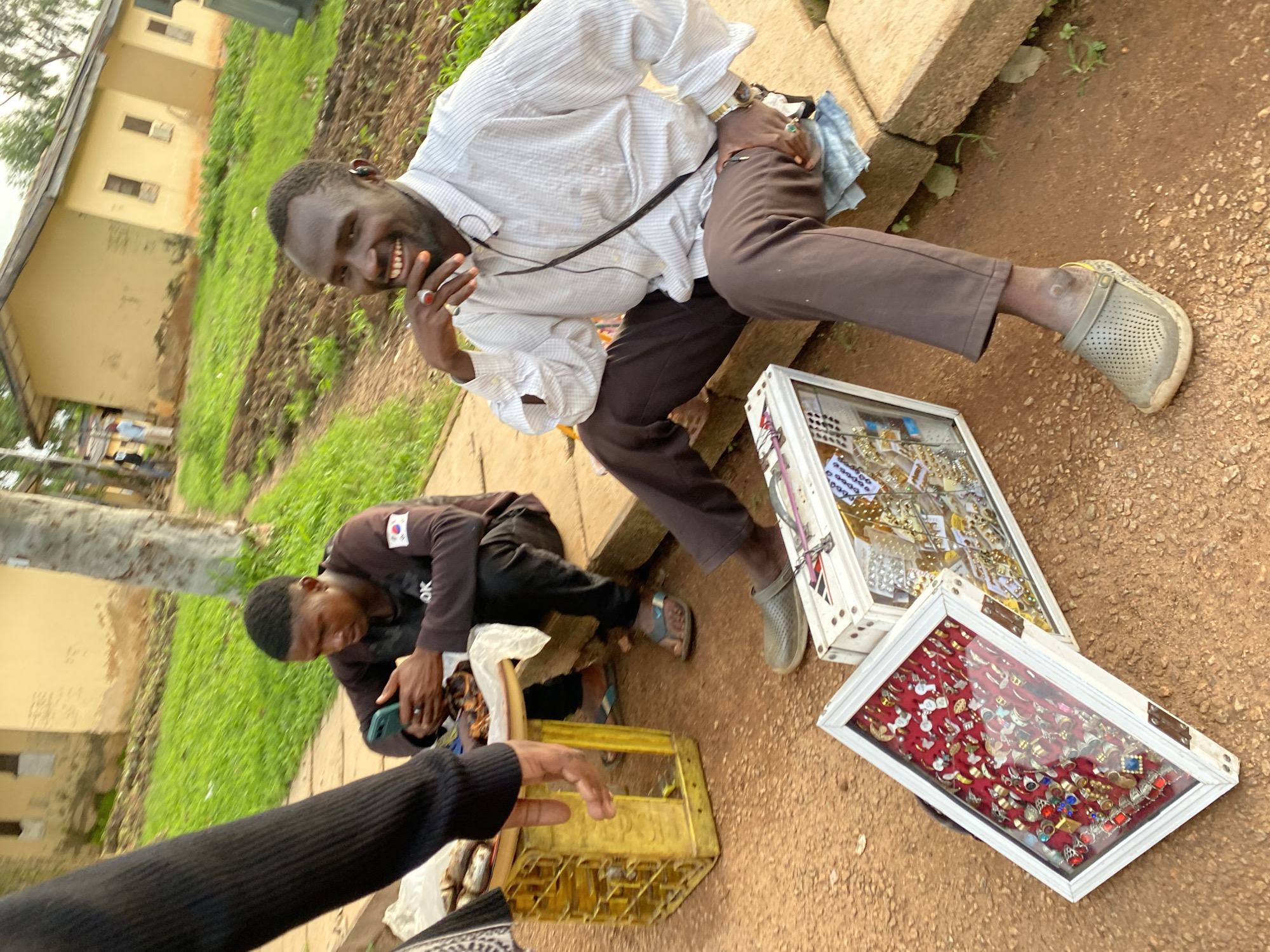 The image depicts an outdoor market scene in Rafin Par, Nigeria, with a focus on two men. The man in the foreground, wearing a light-colored, striped long-sleeved shirt and brown pants, is seated on concrete steps and smiling at the camera. He has a ring on his right index finger and appears to be wearing an earpiece. To his left, another man is seated on a yellow crate, looking down at a smartphone he holds in his hands. He is wearing a dark long-sleeved shirt with a patch on the sleeve that displays the South Korean flag. In front of the seated men, two white display cases filled with assorted rings are laid out on the reddish-brown ground. One case contains rings on a red velvet lining, while the other displays a variety of rings and jewelry pieces. In the background, there are simple, light-colored buildings and overgrown green grass.