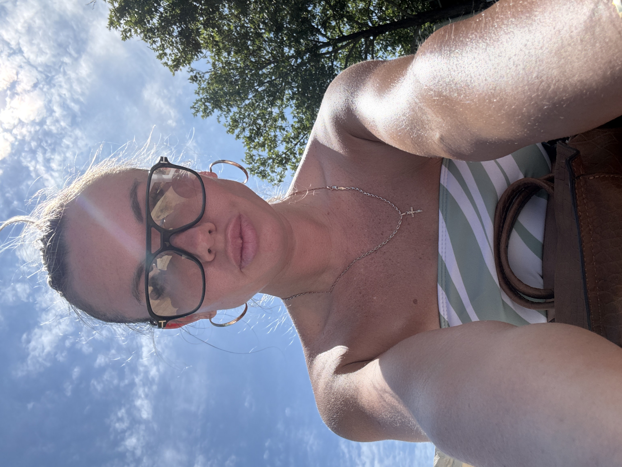 A woman, seen from a low angle, takes a selfie outdoors on a sunny day with a blue sky and scattered clouds. She is wearing large, dark-rimmed glasses and a strapless swimsuit with green and white stripes. A silver chain with a cross pendant is visible around her neck, and she has large hoop earrings. Her hair is pulled up. Her right arm is extended towards the camera, and a portion of a brown handbag is visible at the bottom of the frame. The background includes green trees and a bright sky. The scene appears to be a park or other outdoor recreational area.