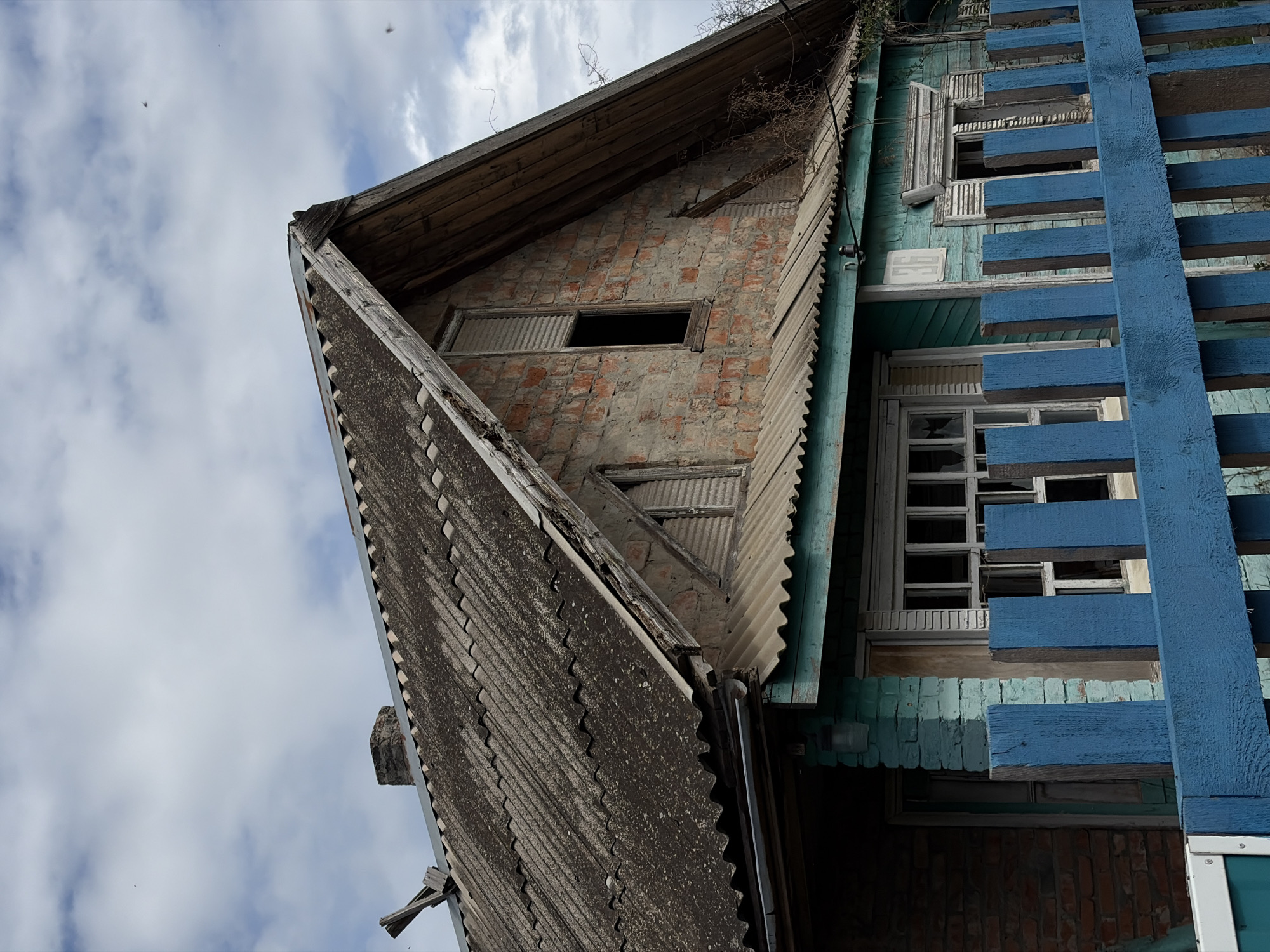 The image shows a dilapidated building, likely located in an urban environment within an unidentified city. The architecture suggests the building is old and in disrepair, evidenced by the weathered brickwork, damaged roofing, and faded paint. The structure appears to be a multi-story building with visible window frames, some broken or missing glass panes. A blue, wooden structure, possibly a fence or railing, is prominently positioned on the right side of the frame, adding a stark contrast to the aged and deteriorated facade of the building. The sky is visible in the background, partially obscured by the building's structure.