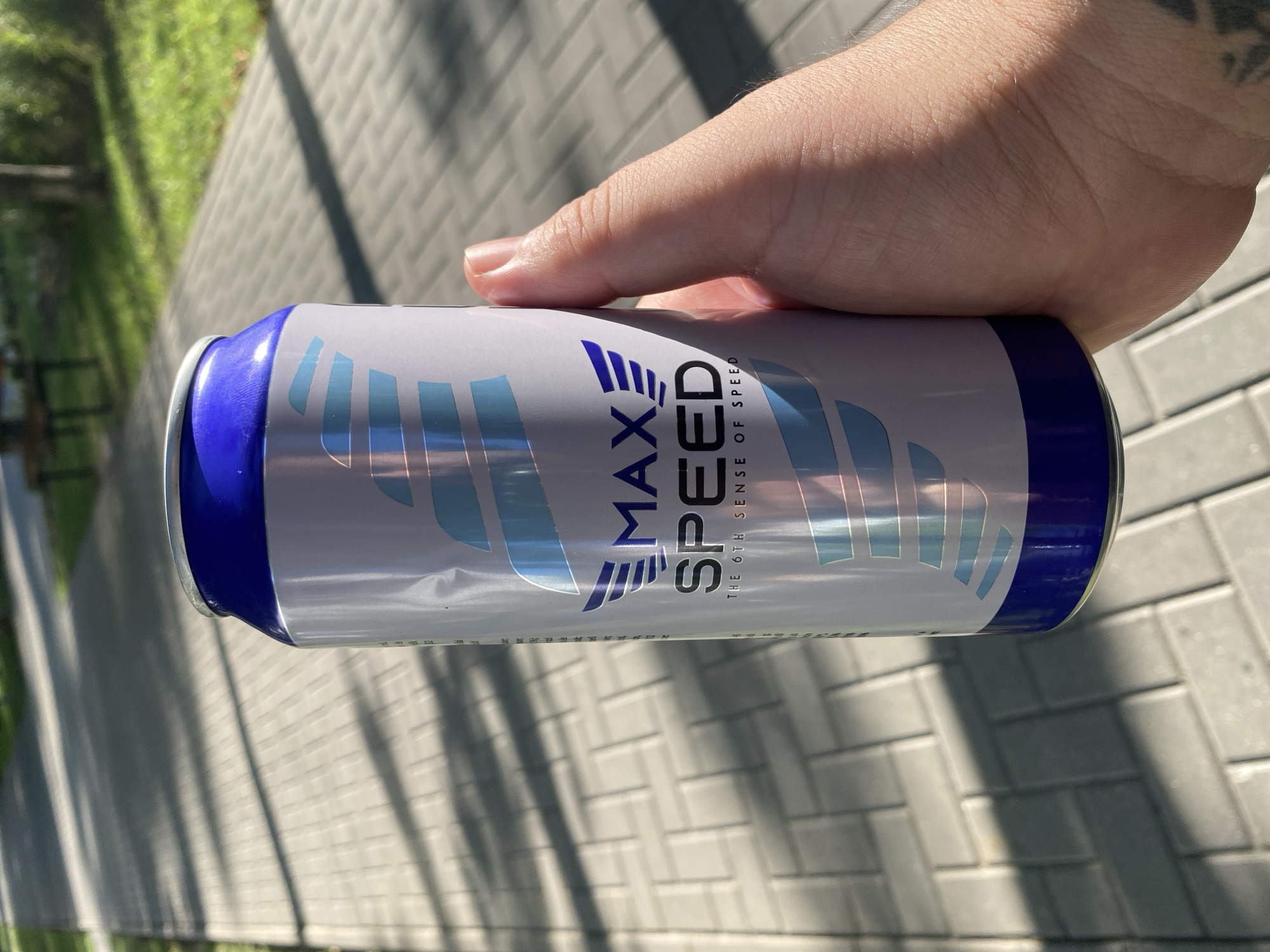 In Chișinău, Moldova, a hand holds a can of "MAX SPEED" energy drink outdoors. The can is predominantly white and silver with blue accents and features stylized wings and text. The background shows a paved walkway with shadows cast by trees, suggesting it is daytime and sunny. A patch of grass is visible to the left of the walkway.