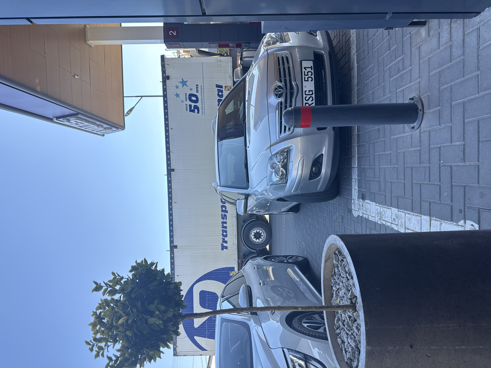In Comrat, Moldova, a gas station canopy casts a shadow over a scene featuring several cars and a large semi-trailer truck. In the foreground, a silver sedan is parked on the patterned grey brick pavement, its license plate reading "RSG 551". To its left, another silver car is partially visible. A concrete planter containing a small tree stands to the left of the sedan. Behind the cars, the white trailer of a semi-truck is emblazoned with a large blue logo and the text "Transport" and "50 ans sur les routes d'Europe". The gas station canopy is supported by white pillars, one of which has a red sign with the number "2". The sky above is clear and blue.