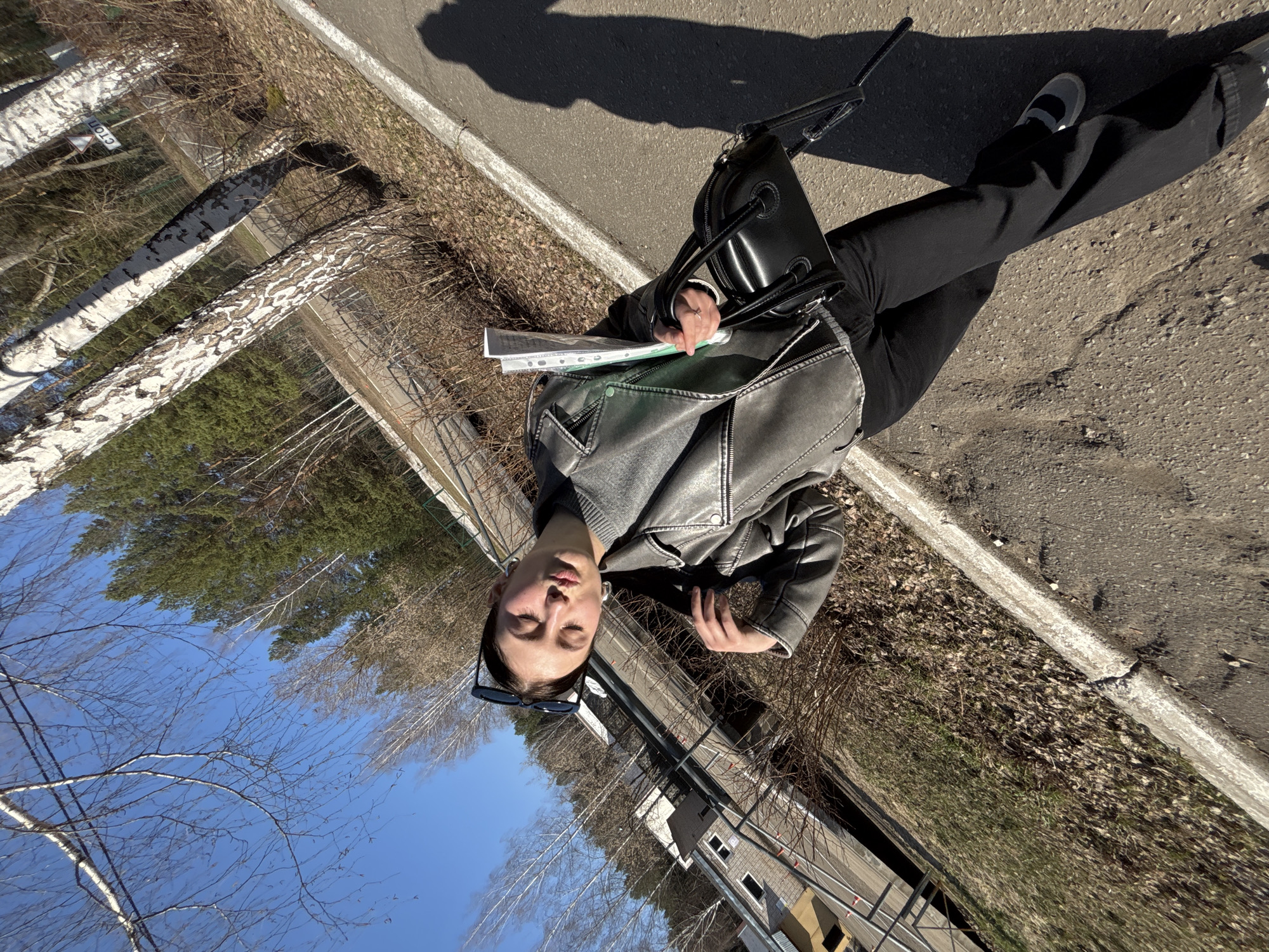 This full-shot outdoor photograph, taken in Chaikovsky, Russia, features a person standing on an asphalt path next to a concrete curb and an area of dried vegetation. The image appears rotated 90 degrees counter-clockwise; when viewed upright, the subject is a young woman positioned towards the right side of the frame, looking slightly towards the left.
The woman is dressed in a dark grey or black leather-like jacket over a grey top, dark wide-leg trousers, and dark flat shoes with a grey vamp and strap across the foot. She carries a black shoulder bag on her left arm, and in her left hand, she holds a clear plastic folder containing papers or documents. Her dark sunglasses are pushed up onto her head. Her eyes are closed or squinted, suggesting she might be facing bright sunlight.
The setting appears to be a roadside or path bordering a natural area, possibly at the edge of a forest or wooded section. The background features numerous trees, including distinctive white-barked birch trees and other bare deciduous trees, indicating late winter or early spring. Further back, evergreen pine trees are visible, along with glimpses of buildings, possibly residential or small industrial structures, behind a fence. The sky is clear and blue.
The time of day is sunny, likely mid-day, as evidenced by the bright light and the distinct, elongated shadows cast by the person and nearby objects onto the asphalt path. These shadows stretch diagonally across the surface. A notable detail confirming the location is a white triangular sign visible behind the birch trees, which bears the Cyrillic word "СТОП" (STOP). The ground cover consists of sparse, dry vegetation and fallen leaves.