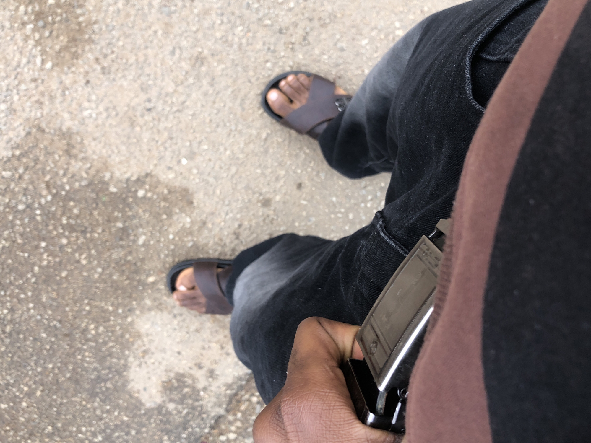 The image shows a close-up, overhead view of a person's legs and hands. The person is wearing dark wash denim jeans and brown leather sandals. Their left hand is visible, holding onto a metal belt buckle. The background is an outdoor ground surface, appearing to be asphalt or concrete with small stones and some lighter colored patches, consistent with a street or sidewalk in Jos, Nigeria.