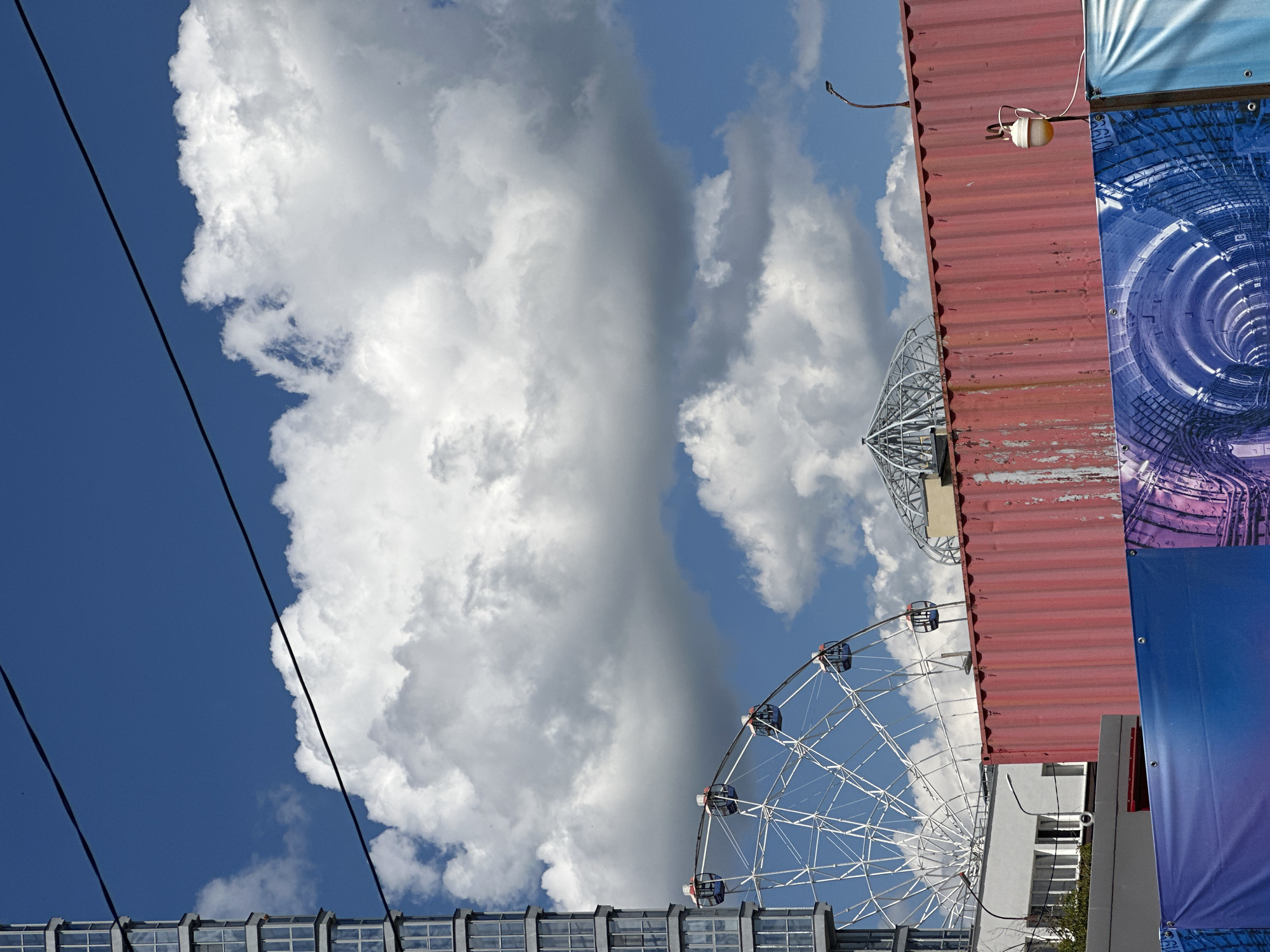 This is a vertical, eye-level, medium shot photograph of a construction site under a bright, cloudy sky. In the foreground, a large, weathered, red corrugated metal shipping container is prominently displayed, positioned horizontally across the frame. Attached to the container, to the right, is a white electrical fixture with a yellow light. Below the container, a large blue banner is visible, featuring a colorful graphic of a tunnel with intricate architectural details and lighting. To the left of the container, a portion of a white, modern building with several windows can be seen.
In the mid-ground, a large, white Ferris wheel with multiple gondolas is partially visible on the left side of the frame. Behind the Ferris wheel and the container, a geodesic dome structure with a metal lattice framework rises against the sky.
In the background, a tall, multi-story building with a grid-like facade of windows is situated on the far left. The sky is a vibrant blue, dotted with large, fluffy white clouds that dominate the upper half of the image. Two black electrical wires are stretched across the top of the frame.
This scene appears to be located in Nizhny Novgorod, Russia, possibly near an entertainment or development area. There are no people visible in the image.