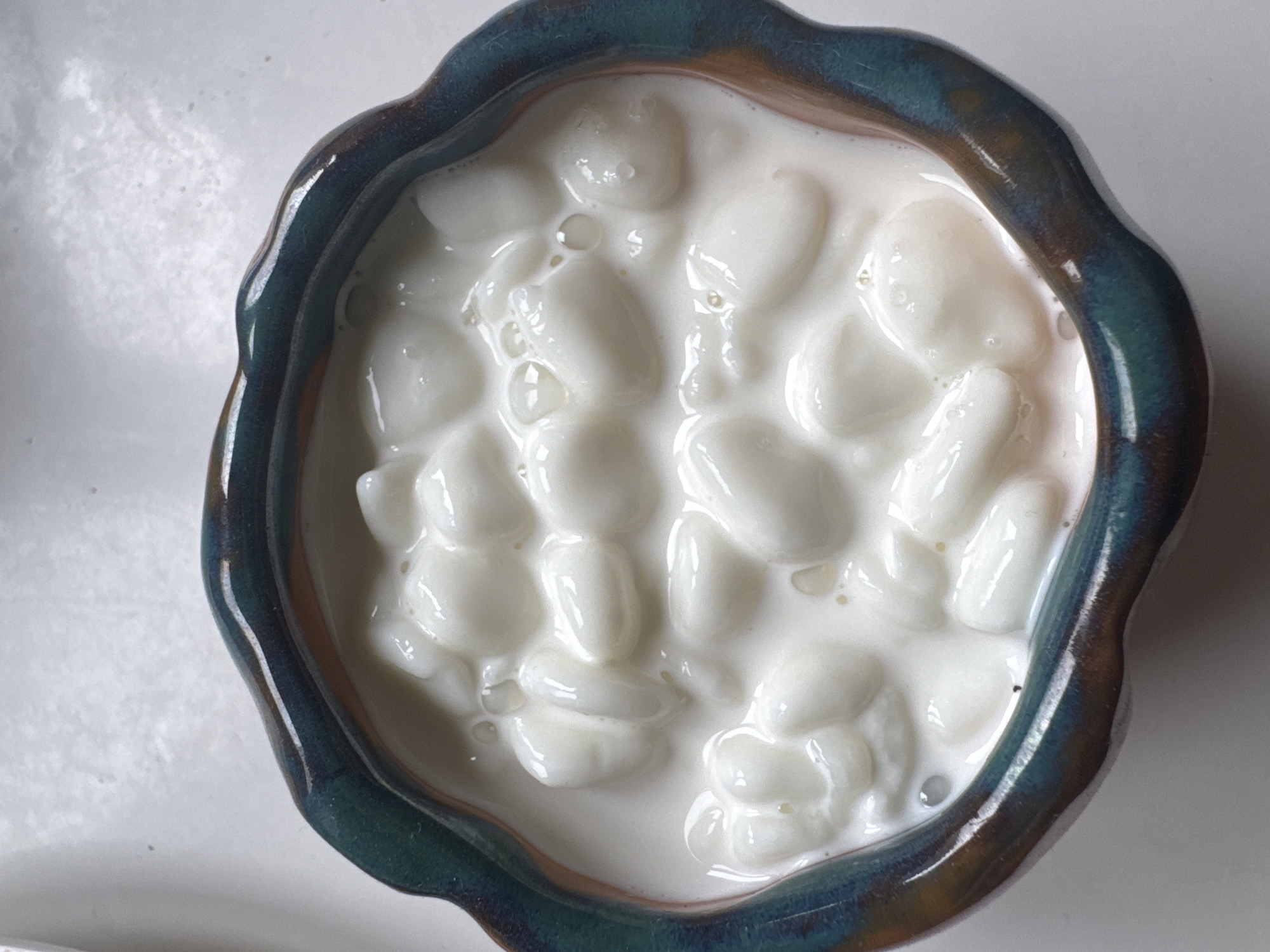 An overhead, close-up shot features a small, irregularly shaped ceramic bowl filled with white, oval-shaped food items suspended in a creamy white liquid. The individual food pieces are smooth, rounded, and appear soft, possibly resembling dumplings or gnocchi. Small bubbles are visible on the surface of the liquid.
The bowl's rim has a distinctive, wavy edge, finished with a glossy, variegated glaze. This glaze transitions from a deep blue-green to mottled brownish-gold hues, indicative of a reactive or handcrafted finish. The interior of the bowl is obscured by its contents. The background is a plain, light-colored surface, likely a table or countertop.
The image is a still life, with no activity or individuals present. There are no visual cues to suggest a specific time of day, weather, or a particular location, including Moscow, Russia. No text is visible anywhere in the image.