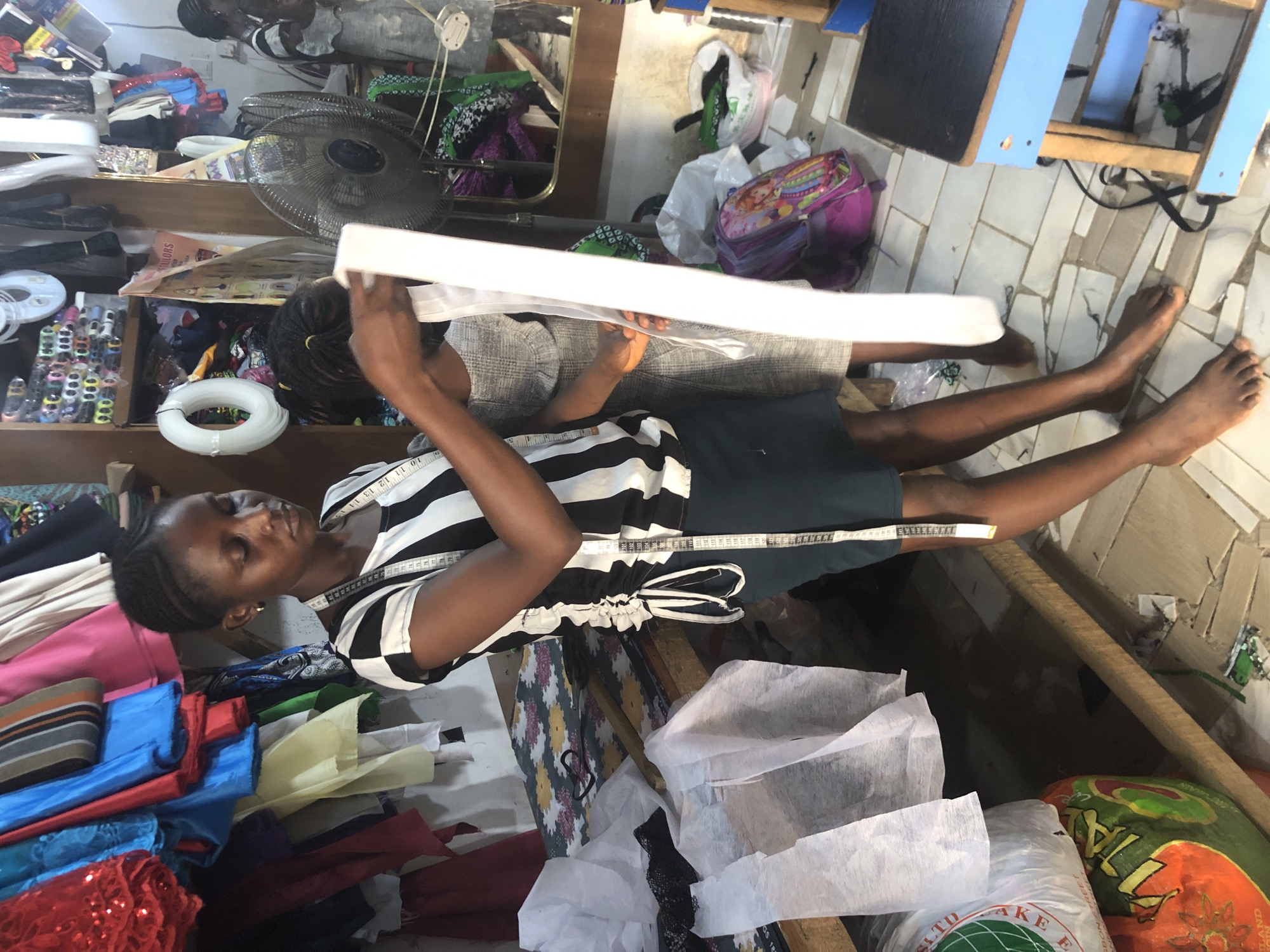 In a dimly lit, cluttered tailor shop in Ado, Nigeria, two young girls are engaged in their work. The older girl, with her hair in braids, wears a black and white striped shirt and a dark skirt, a measuring tape draped around her neck. She holds a long strip of white fabric, her fingers delicately manipulating it. Beside her, a younger girl in a light-colored dress stands, observing or perhaps assisting. The shop is filled with bolts of colorful fabric, spools of thread, and other sewing supplies. A fan is visible in the background, a welcome sight in the Nigerian heat. The floor is tiled, and a small stool with a blue frame and black top sits in the foreground, possibly for the tailor's use. Bags and personal belongings are scattered around, adding to the shop's lived-in feel.