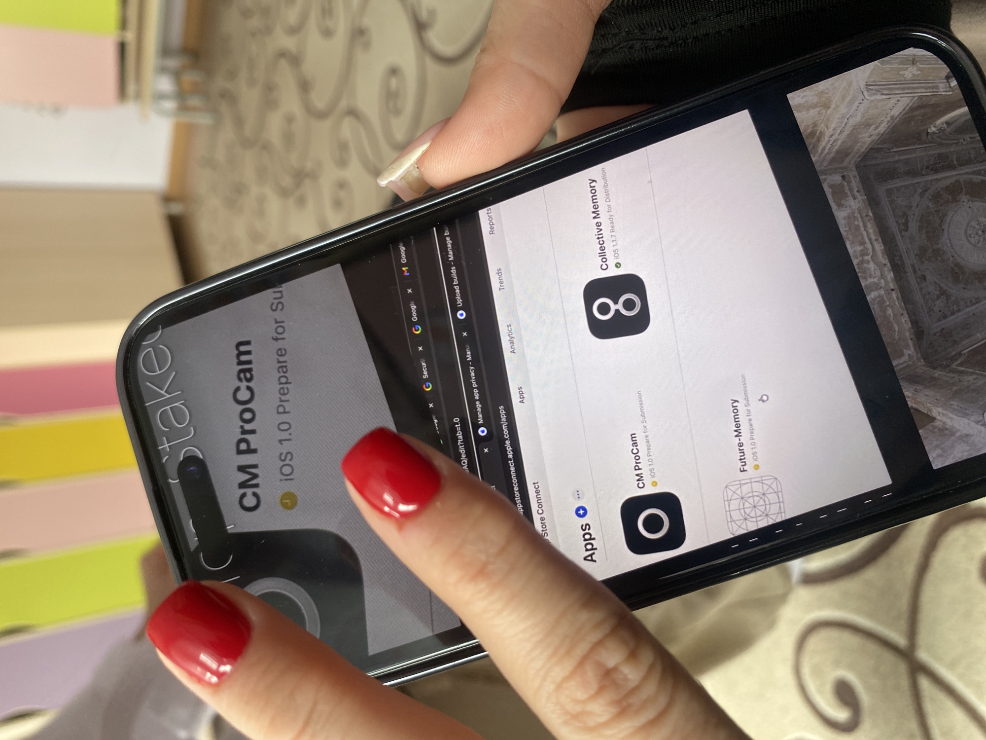 A close-up, angled shot shows a person's hands interacting with a black-cased smartphone in an indoor setting. The phone displays what appears to be an Apple App Store Connect interface. The user's left hand, with red polished nails, has a finger poised over the screen, while part of their right hand, with natural nails, is visible holding the device.
On the phone's screen, multiple browser tabs are visible on the left, including "G Google", "G Securit", "Manage app privacy", and "Upload builds". The main section of the screen is focused on app management within "appstoreconnect.apple.com/apps". Prominently displayed are three app entries: "Collective Memory" (iOS 1.1.7 Ready for Distribution, with an infinity-like icon), "CM ProCam" (iOS 1.0 Prepare for Submission, with a camera icon), and "Future-Memory" (iOS 1.0 Prepare for Submission, with a geometric icon). Headers like "Apps", "Analytics", "Trends", and "Reports" are also visible. A partial reflection on the screen shows a detailed, ornate ceiling, possibly part of a building's interior. Text like "Stake" is partially visible near the top left of the phone's display, likely part of a phone widget or status.
The background reveals a light-colored wall with a subtle, swirling patterned wallpaper. To the left, colorful stacked objects or shelves in shades of pink, green, and yellow are blurred. The scene suggests an individual working on app development or management within a personal or office environment in Chaikovsky, Russia.