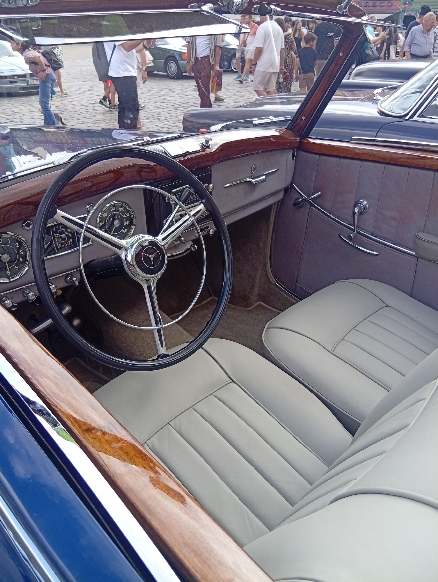 The image captures the interior of a classic Mercedes-Benz convertible, viewed from the driver's side. The car's interior features light-colored leather seats, a wooden dashboard, and a chrome steering wheel with the Mercedes-Benz emblem. A clear reflection in the rearview mirror shows a street scene with several people walking around. One person in the reflection is wearing a pink shirt and blue jeans, and another is wearing a white t-shirt and black pants. Other individuals are visible in the background, dressed in casual attire. The setting appears to be an outdoor event or gathering, possibly a car show, given the presence of multiple vehicles and a crowd. The architecture in the background is indistinct, but the overall ambiance suggests an urban environment. The text visible in the reflection on a building in the distance reads "TRECZO", which is a brand. Based on the metadata of the image, the location is identified as Wrocław, Poland. The image is taken in daylight, with the sun casting light and reflections on the car's surfaces and the surrounding environment.