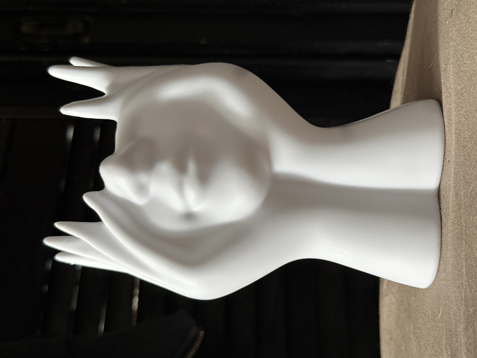 A white ceramic vase, shaped like a stylized human face with hands cupped around it, is centered in the frame. The face has closed eyes, a subtle smile, and the hands form a crown-like shape above the head. The vase rests on a textured, tan surface, possibly a piece of furniture. The background is dark and out of focus, with horizontal lines suggesting blinds or a slatted surface. There is no visible indication of this scene being located in Moscow, Russia, or any other specific geographic location.