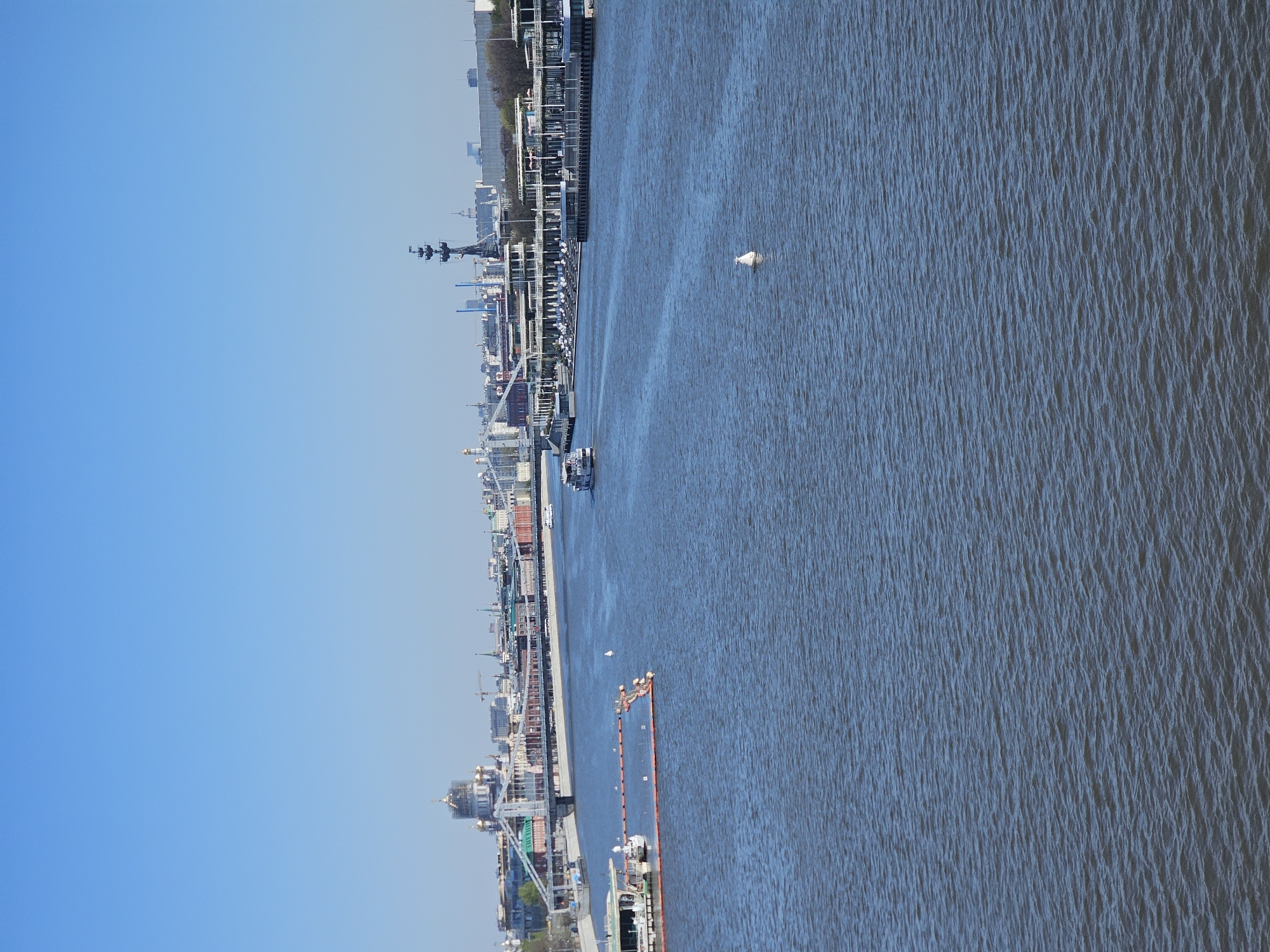 The image depicts a section of the Moskva River in Moscow, Russia. The view is likely taken from a high vantage point, possibly a bridge or elevated building. The river flows from the upper-right to the lower-left of the frame. The right side of the image is dominated by the river’s surface, showing a textured pattern of water. A white floating object, possibly a buoy or debris, is visible in the river.
The left side of the image shows the riverbank with a row of buildings and structures along it. There are modern buildings, historical structures, and industrial elements. A naval ship is visible along with numerous cranes along the waterfront.