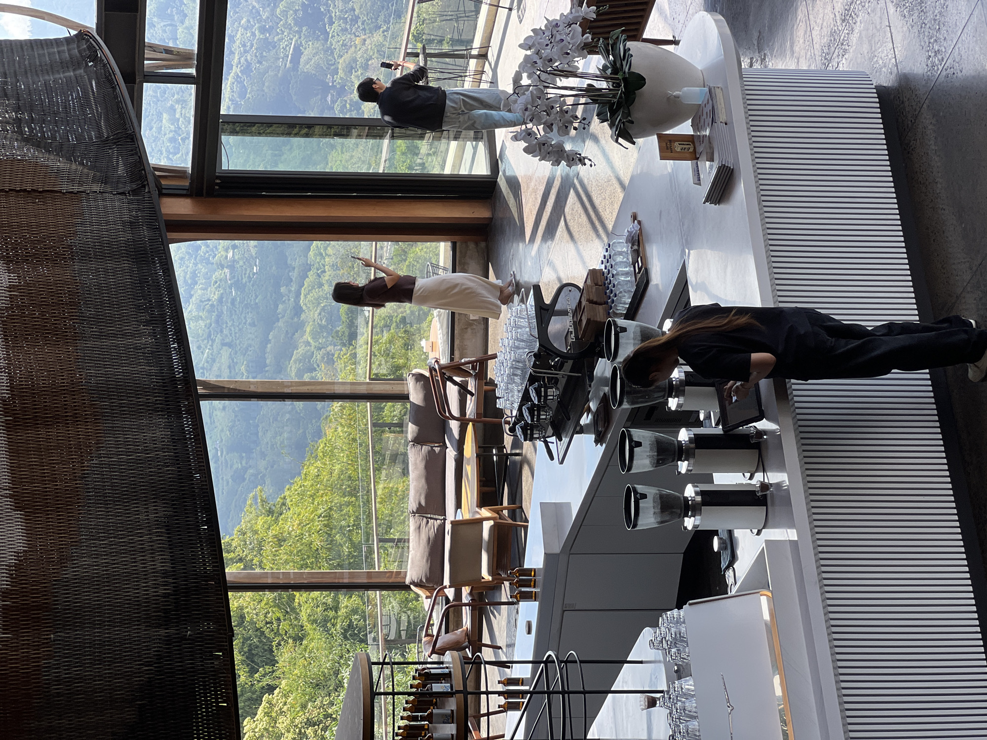 This image captures the interior of a modern cafe with a scenic view of lush green mountains in Kintamani, Indonesia. In the foreground, a sleek white counter is visible, equipped with multiple coffee grinders and a barista station. A woman in black attire is attending to the counter, interacting with a tablet device. Behind the counter, a white orchid plant in a large pot adds a touch of elegance. Through the large glass windows, a breathtaking panorama of rolling hills and dense vegetation unfolds, indicative of the natural beauty of the Kintamani region. The cafe's architecture features a high ceiling with a woven dark-colored canopy, and a comfortable seating area with a sofa and chairs is visible in the mid-ground. Two patrons are present, a woman standing near the window holding a phone, and a man further back, also with a phone in hand, appearing to take in the view. The overall ambiance suggests a tranquil and aesthetically pleasing environment, blending modern design with the stunning natural landscape.