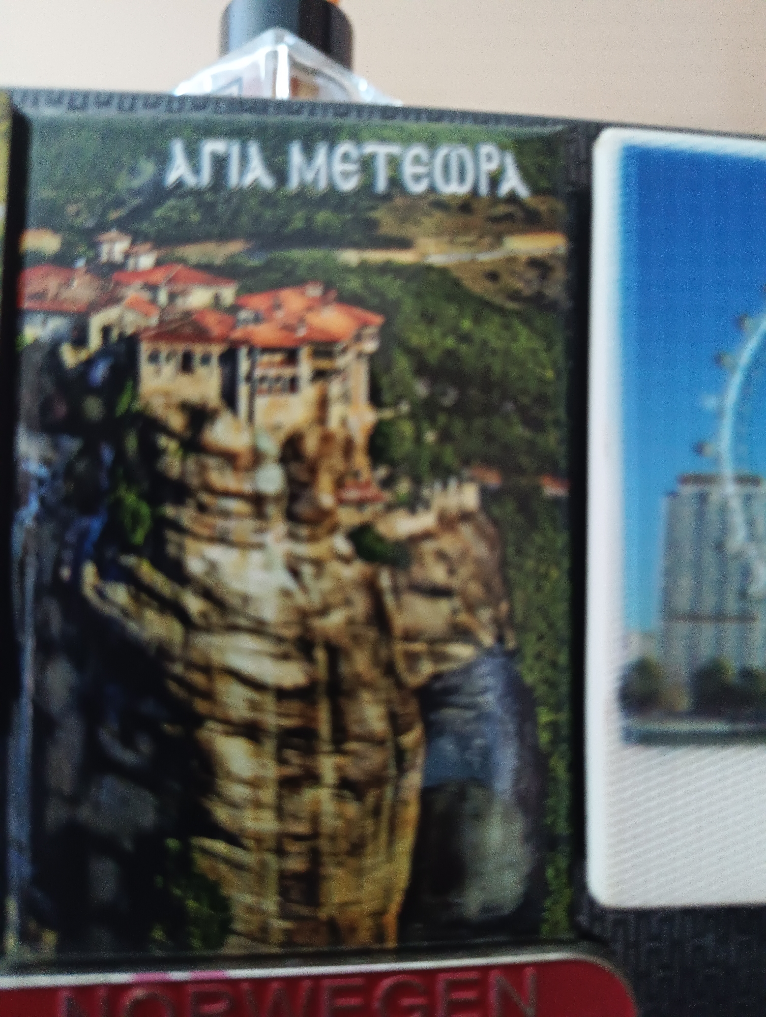The image shows a close-up view of two souvenirs. The one on the left features a postcard-like image of monasteries built on top of rock pillars, with Greek lettering above. The one on the right shows a Ferris wheel in a city with a blue sky. At the bottom of the image, red text that reads "NORWEGEN" is visible. The scene is likely a collection of souvenirs from different locations, and the reference to Karnobat, Bulgaria, appears to be an incorrect assumption as there are no visual cues in the image to suggest this location.