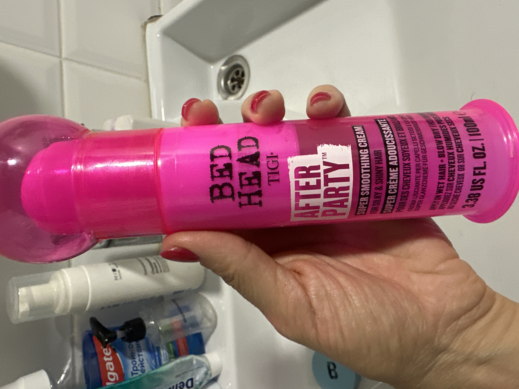 A close-up shot shows a hand holding a bright pink Bed Head "After Party" hair smoothing cream bottle. The bottle has black text and a clear plastic cap. The background is a white tiled wall and a white sink with a silver drain. The hand has red nail polish. There are other toiletries on the sink counter including a tube of Colgate toothpaste and a white pump bottle. The scene is indoors and appears to be a bathroom. The image location is not discernible from the visual information provided.