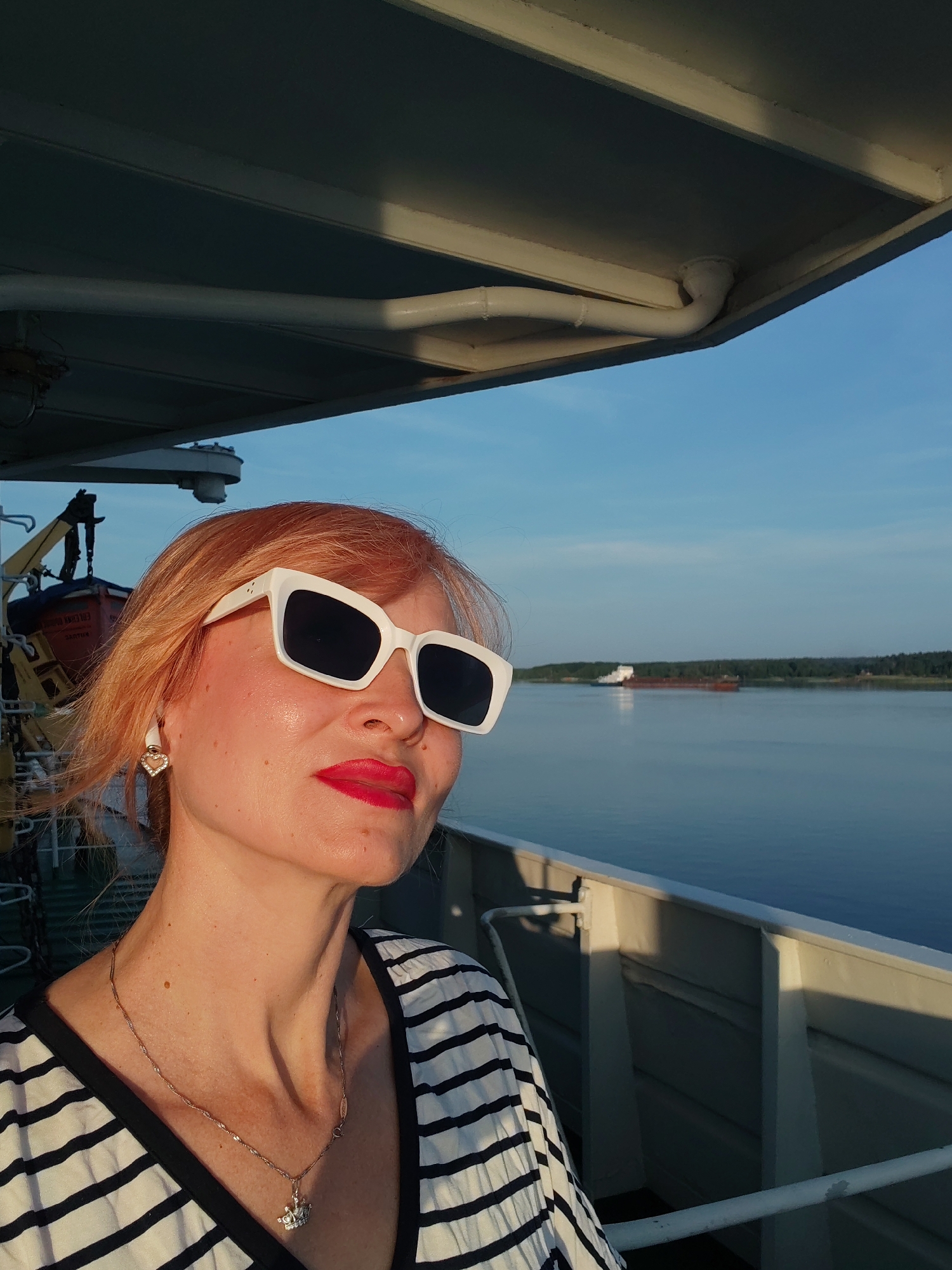 A medium shot captures a woman with reddish-blonde hair and bright red lipstick posing for a selfie on a boat. She is wearing large white cat-eye sunglasses and a black and white striped shirt. A heart-shaped earring with small white stones adorns her left ear, and a silver necklace with a crown pendant rests on her collarbone. The background shows a calm body of water with a distant cargo ship. The sky is clear and blue, suggesting it is daytime. The boat's structure, including a portion of its roof and railing, is visible in the foreground. The location is identified as Shchetinino, Russia.