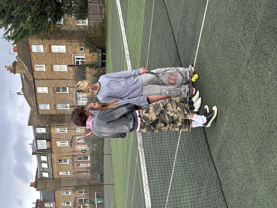 Two people lying on a tennis court, facing each other. One person is wearing a gray and white striped shirt and gray pants, holding a tennis racket. Two tennis balls rest beside them. The second person wears a gray jacket with a pink collar and camouflage shorts, also holding a racket. A tennis net stretches across the frame. Buildings are visible in the background under a cloudy sky.