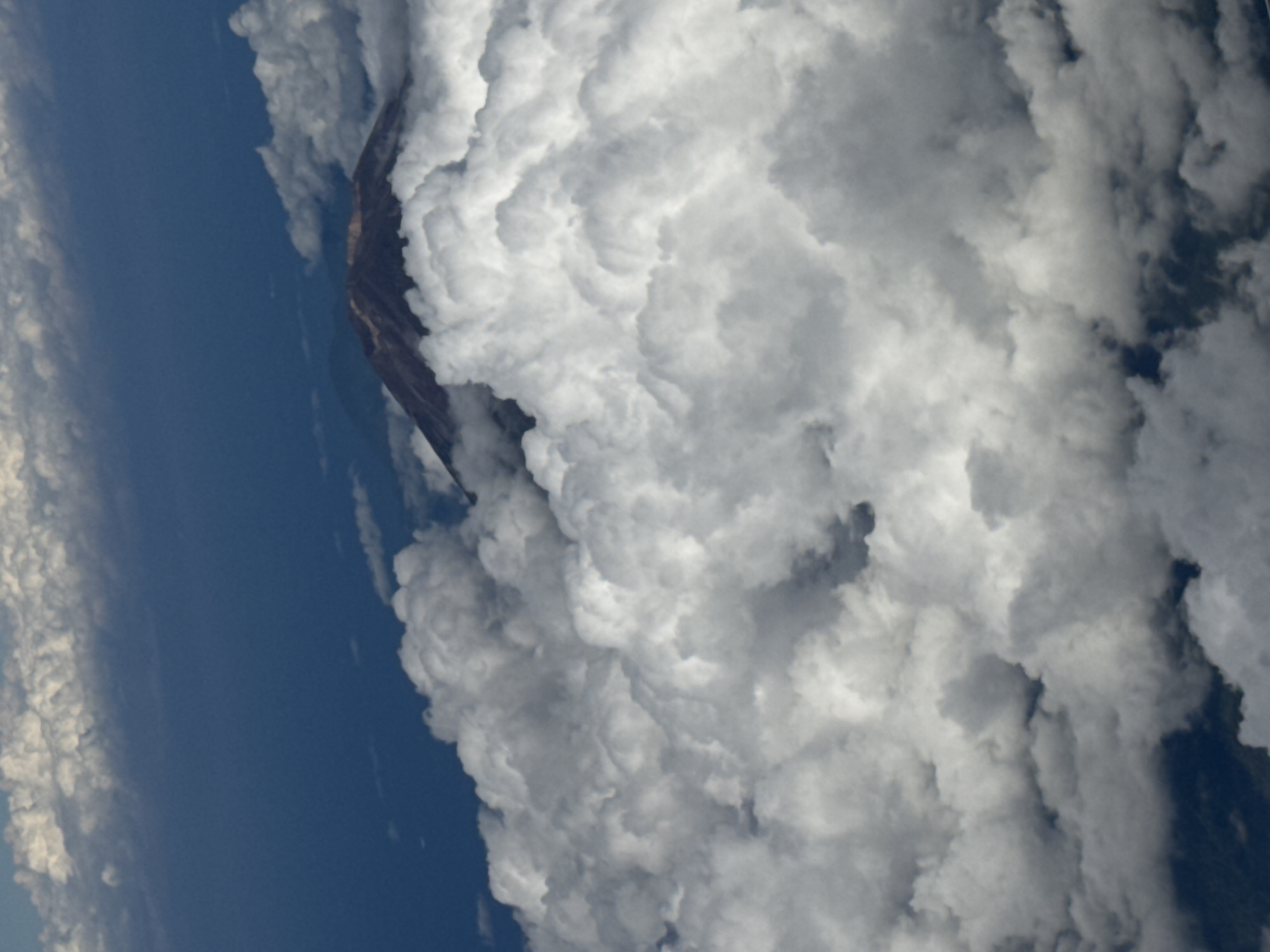 The image, rotated for perspective, depicts a landscape featuring a mountain peak partially obscured by a dense cloud formation. A section of blue sky is visible on the left side of the image, indicating the atmosphere beyond the cloud cover. The clouds are textured with varying shades of white and gray. The mountain peak is dark in color. The location is implied to be in the city of Bangli, Indonesia, although this information is not visually evident in the image itself.