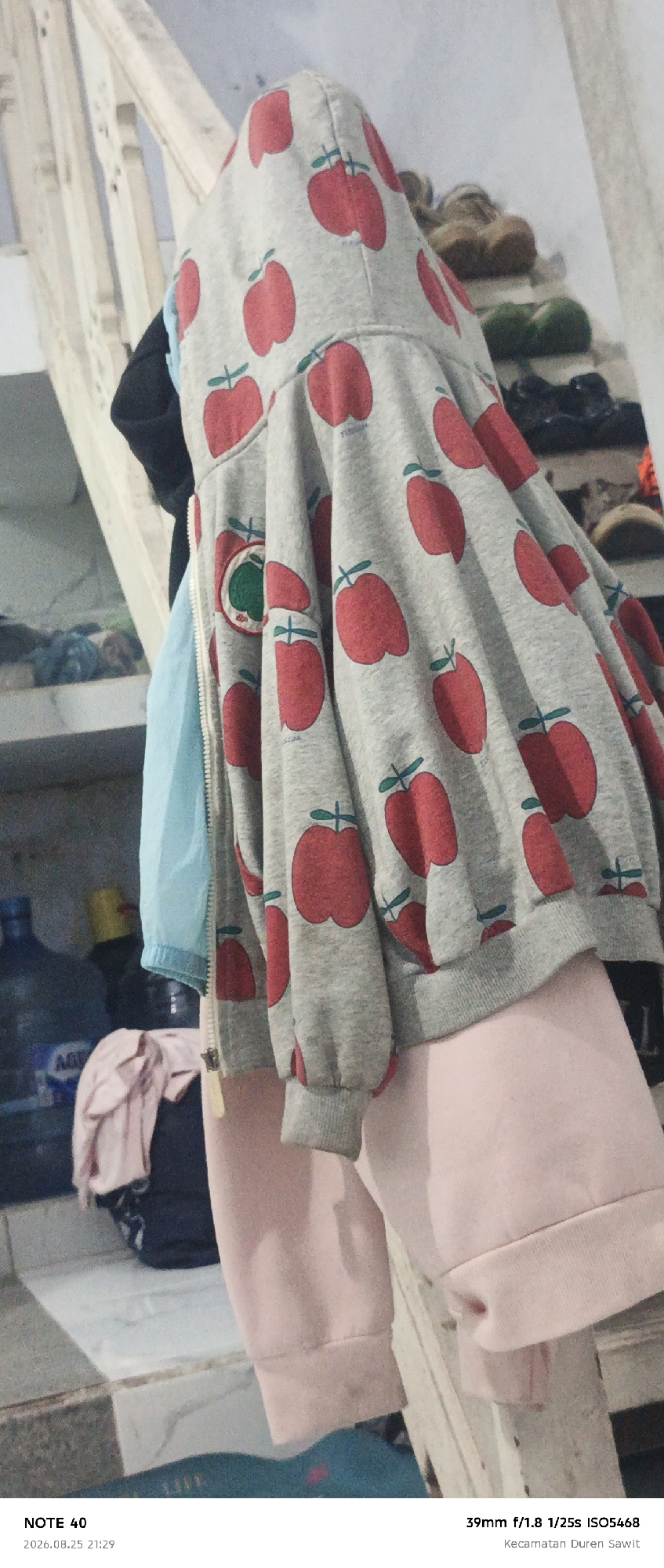 The image shows a close-up of a light gray hooded sweatshirt with a repeating red apple pattern, hanging on a wooden staircase. The sweatshirt is partially obscuring a light blue garment and pink sweatpants. In the background, shelves are visible with various items, including water bottles and shoes. The bottom of the image contains text overlay indicating "NOTE 40" and a date and time of "2026.08.25 21:29", along with camera specifications "39mm f/1.8 1/25s ISO5468" and "Kecamatan Duren Sawit", which is a subdistrict in East Jakarta, Indonesia.