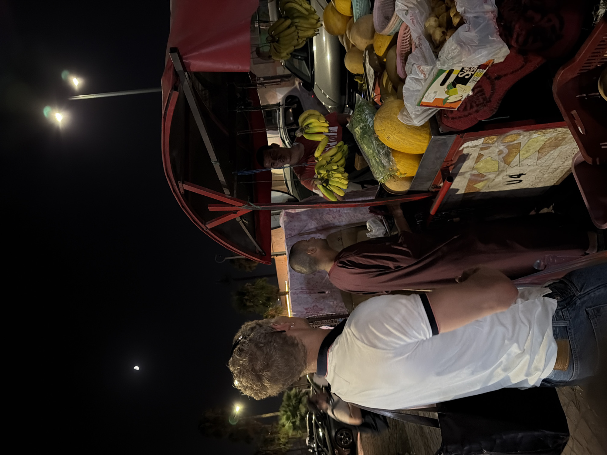 An exterior shot taken at night in Marrakesh, Morocco, depicts a bustling street scene with a fruit vendor and several people. The vendor, a man wearing a red t-shirt, stands behind a cart laden with produce, including a large quantity of yellow bananas hanging from the canopy and in bunches. Melons, some in shades of yellow, are also visible on the cart, along with greenery. A customer, a man with blonde curly hair wearing a white polo shirt and blue jeans, stands facing the vendor, appearing to be in the process of a transaction. Another person, dressed in a maroon robe, stands near the customer, looking towards the vendor. The background reveals palm trees silhouetted against a dark sky where a crescent moon and streetlights are visible. Cars are parked in the background, suggesting an urban environment. The overall impression is that of a vibrant night market in Marrakesh.