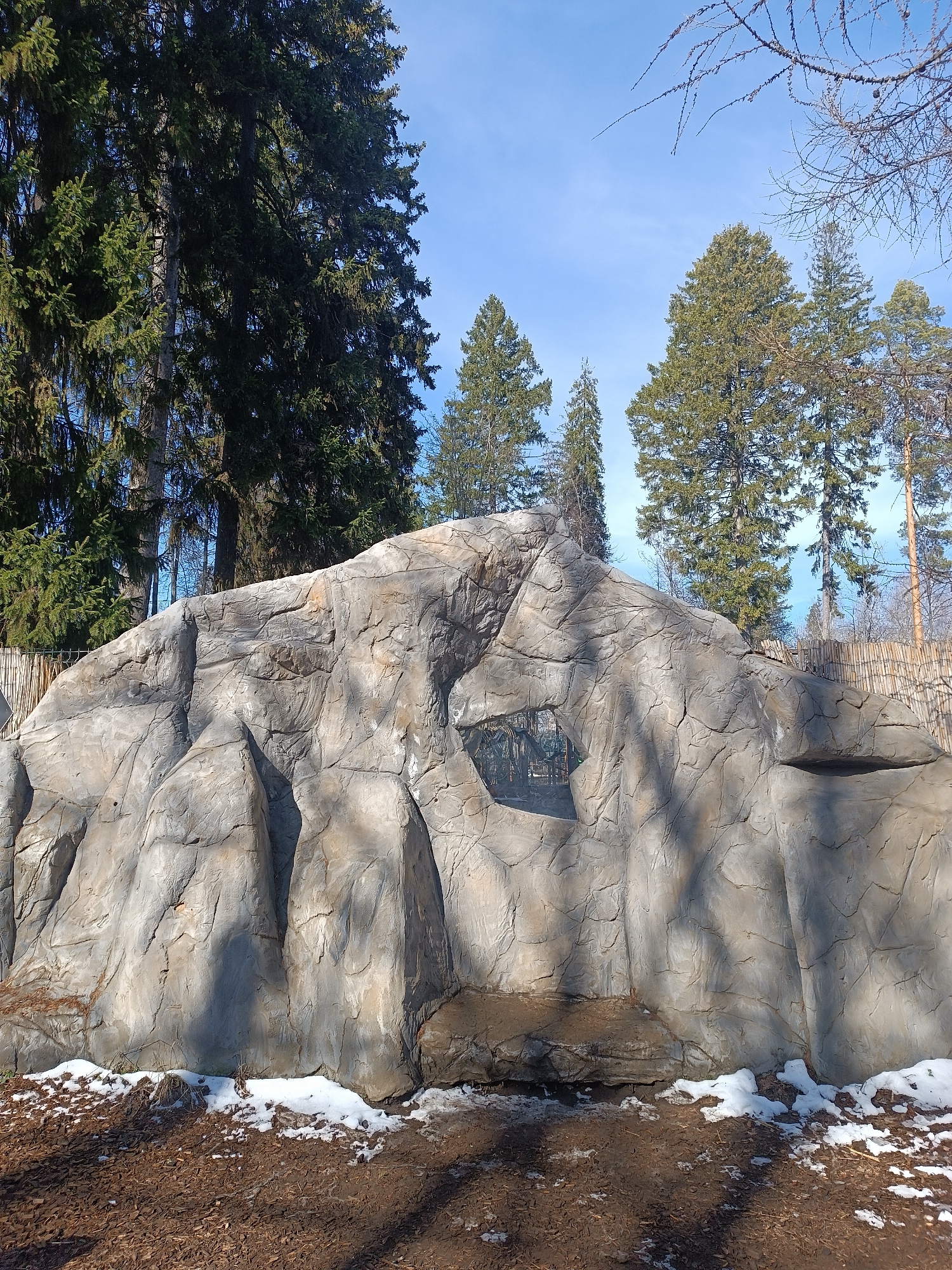 The image shows a large, artificial rock formation in what appears to be a park or zoo setting. The rock is gray with textured surfaces resembling natural stone. There's a small, rectangular inset within the rock, possibly a display or plaque. Patches of snow are visible at the base of the rock, mixed with mulch or wood chips. Behind the rock formation are tall evergreen trees and a section of a bamboo fence. The sky is clear and blue. The specific location is Izhevsk, Russia.