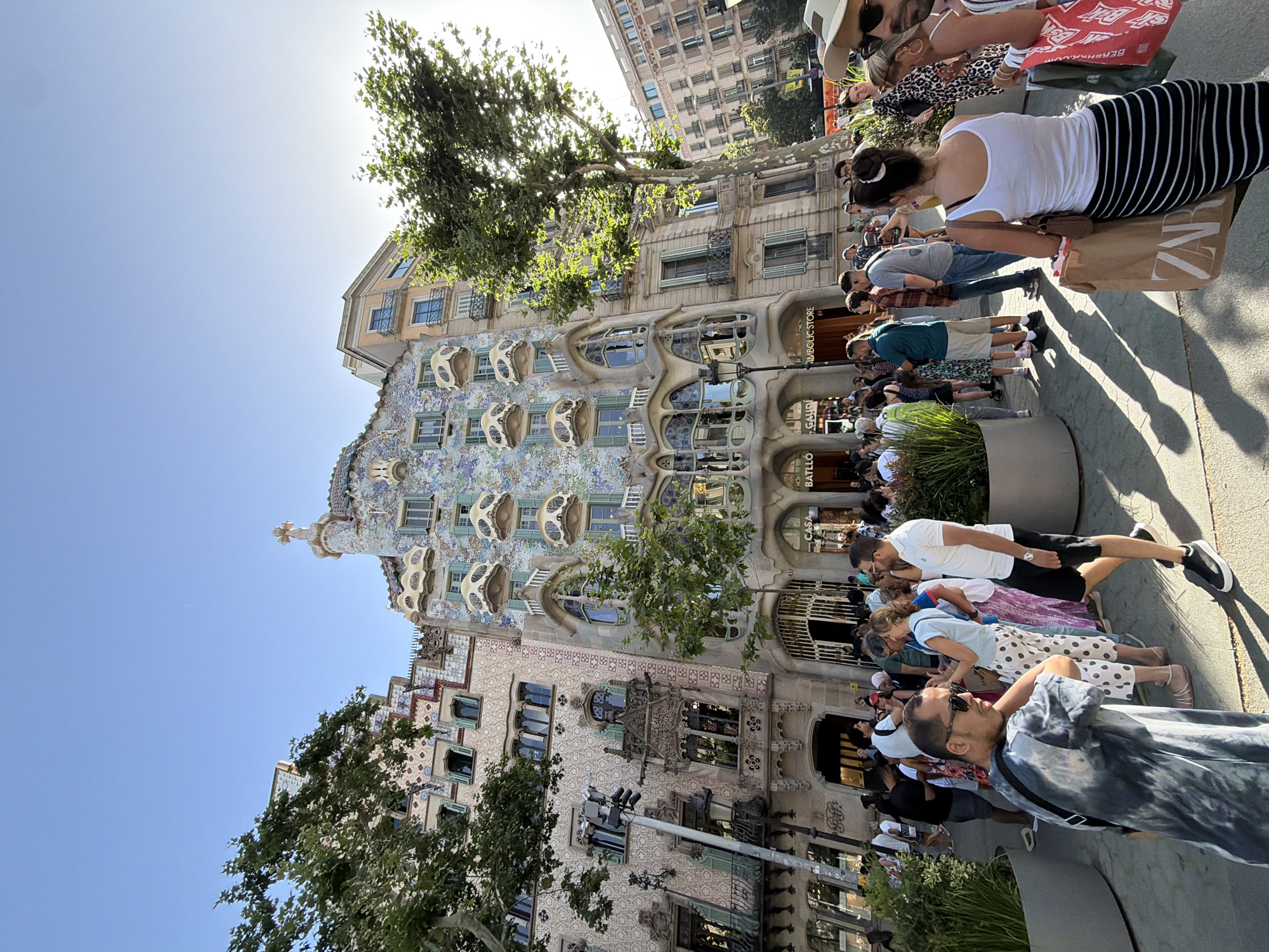 The image shows a street scene in Barcelona, Spain, in front of Casa Batlló. The iconic modernist building, designed by Antoni Gaudí, dominates the background with its distinctive facade covered in colorful mosaics and bone-like balconies. A crowd of tourists and locals is gathered on the sidewalk, some posing for photos, others looking at the building or shopping bags. The sun is shining, casting long shadows on the ground. The atmosphere appears lively and vibrant.