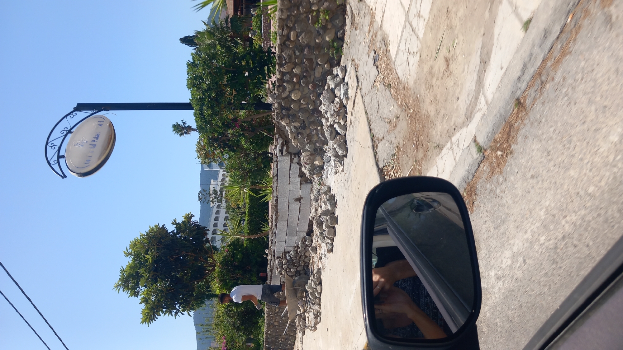 In the city of Heraklion, Greece, on a bright, sunny day, a construction site is visible by the side of a road. A man in a white t-shirt and shorts is working near a wheelbarrow and a pile of stones. Part of a stone wall is under construction, with some sections made of large stones and others of concrete blocks. Debris, including scattered rocks and broken concrete, litters the ground. In the foreground, a car's side mirror reflects a person's hands and arms, suggesting the photo was taken from inside a vehicle. A sign with text, possibly indicating a business name like "Crystal Palace," is suspended above the scene. Lush green trees and foliage surround the construction area, and a white multi-story building can be seen in the background, nestled against a hillside. The overall scene depicts ongoing renovation or construction work in a semi-urban environment.