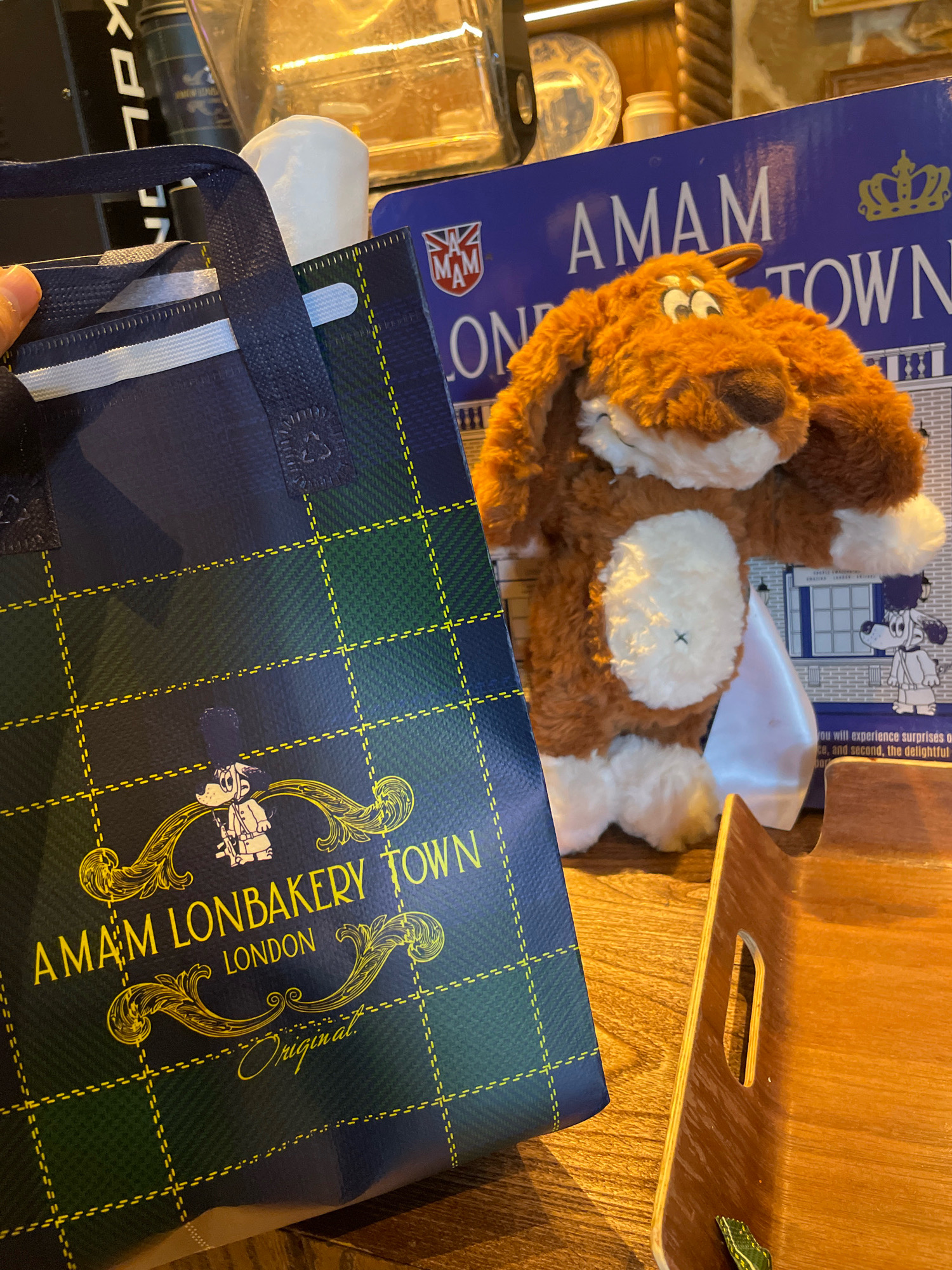 A plush dog toy with brown fur and large, expressive eyes sits on a wooden surface next to a dark blue plaid shopping bag. The bag features yellow stitching and text that reads "AMAM LONBAKERY TOWN LONDON Original." A blue cardboard box with the text "AMAM TOWN" is visible behind the toy and bag. The scene appears to be indoors, possibly in a shop or cafe, with warm lighting and a rustic wooden table. The background includes other objects and decorations that suggest a cozy, eclectic atmosphere. The overall impression is one of a retail or hospitality setting.