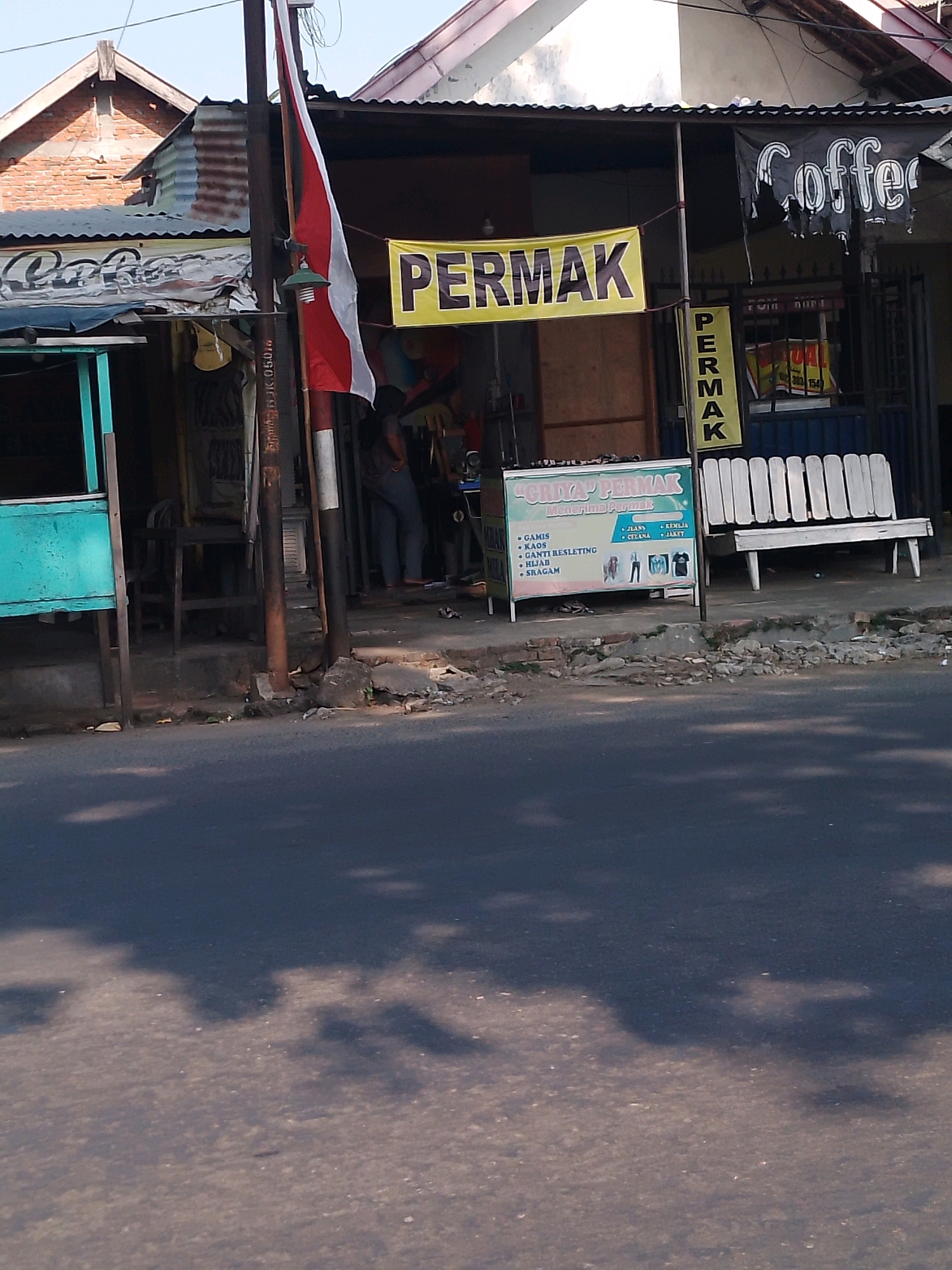 The image shows a street scene in Kertosono, Indonesia. In the foreground, a dark asphalt road is visible with shadows cast by trees or structures. The background depicts a row of shops and stalls along the roadside. A prominent yellow banner with black lettering reads "PERMAK," indicating a tailor or alteration shop. To the right of this banner, another vertical banner also reads "PERMAK." A black banner with "Coffee" is partially visible on a shop to the far right. A white wooden bench is situated in front of one of the establishments. A sign board with text in Indonesian, including "GRIYA PERMAK," "MENERIMA PERMAK," and a list of services like "GAMIS," "KAOS," and "JEANS," is displayed on a stand. A person in dark clothing is standing near the shops. The overall scene suggests a commercial area on a sunny day.